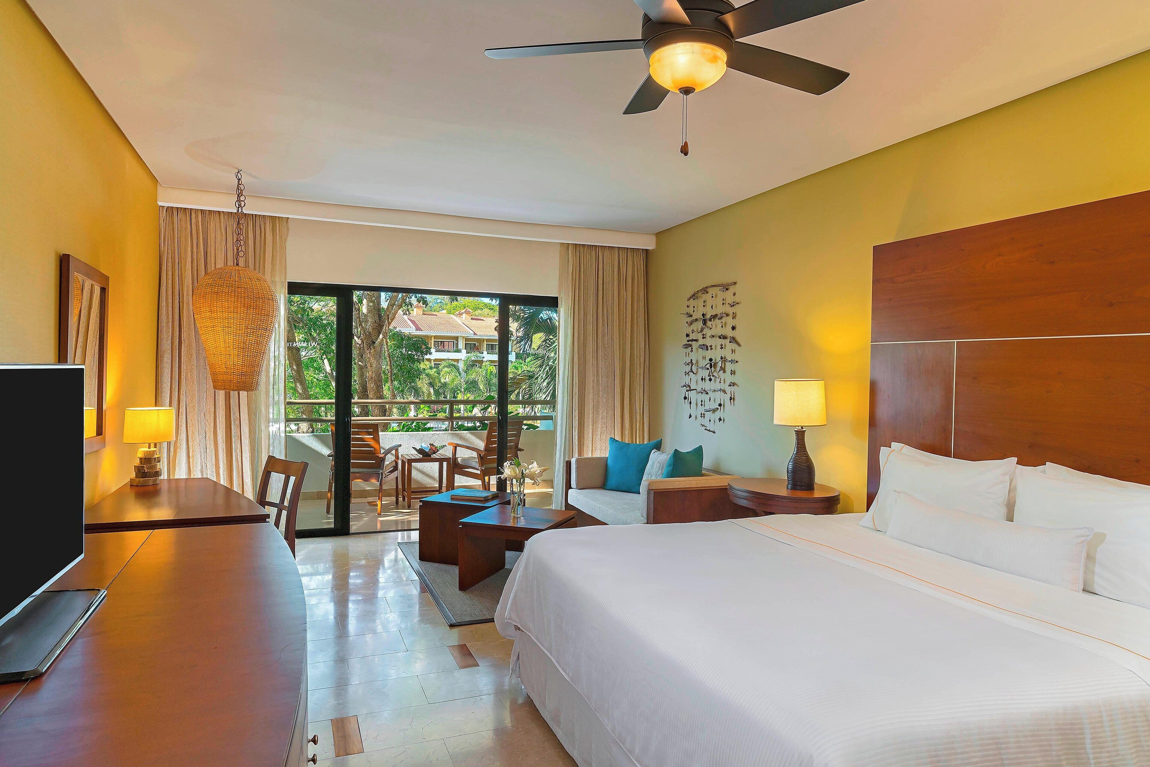 A hotel room with a bed, a desk, a chair, a table, a ceiling fan, and a view of the outdoors.