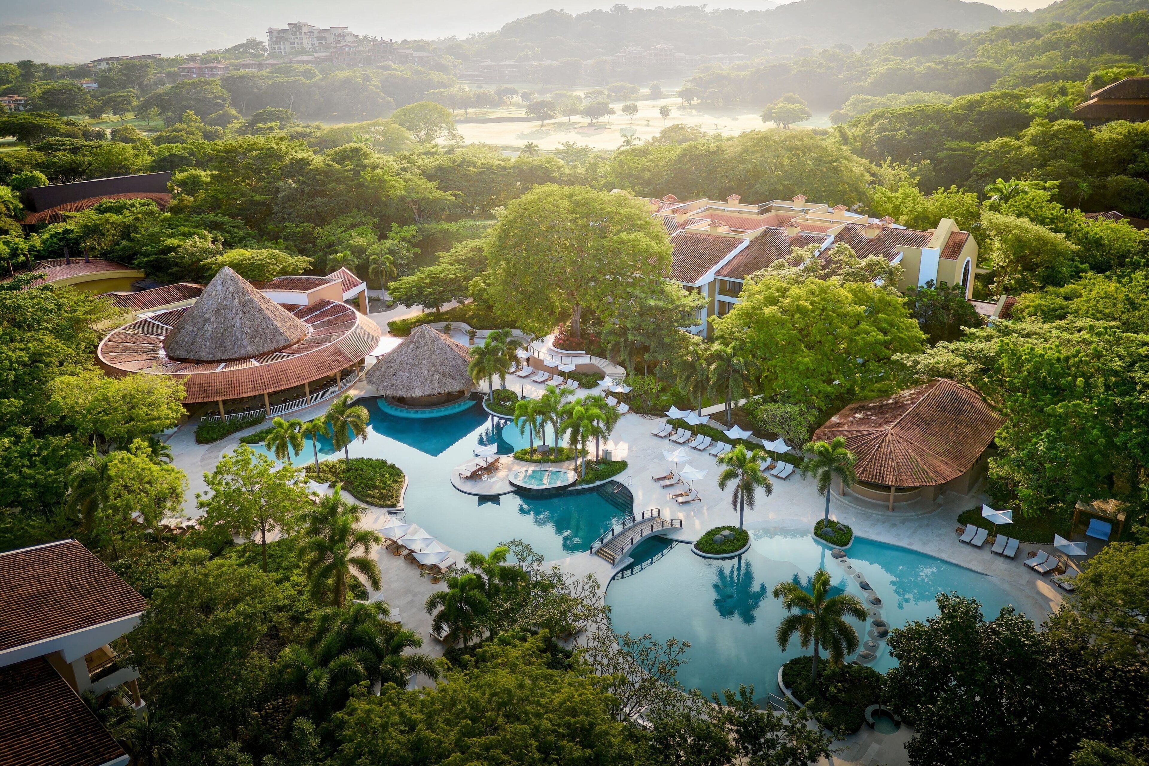 Resort com várias piscinas, palmeiras e construções com telhado de palha.