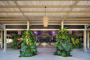 A covered outdoor dining area with tables and chairs, surrounded by large plants and a hanging light fixture.
