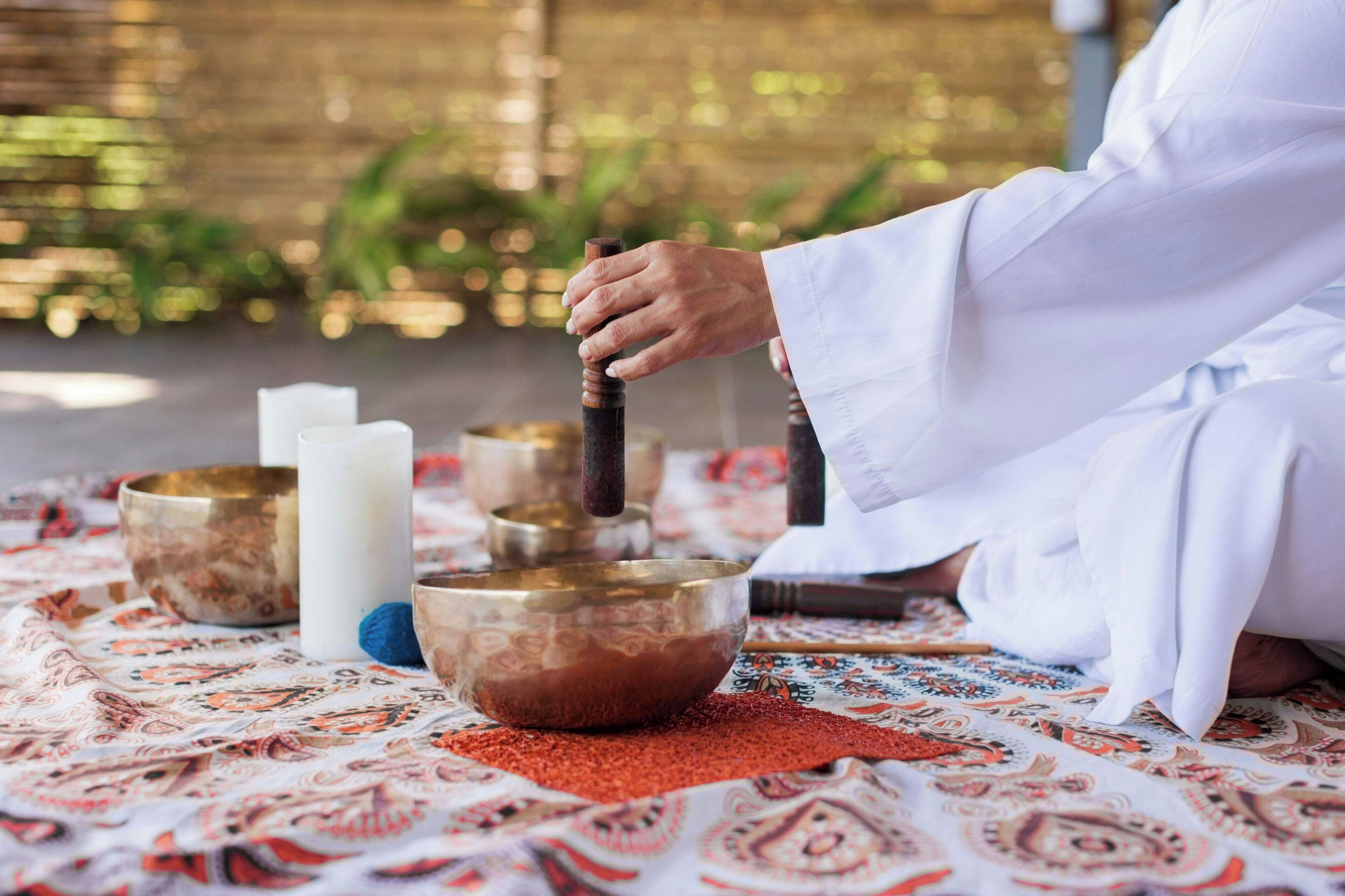 A person sitting on a patterned mat with a meditation bowl, candles, and a wooden mallet.