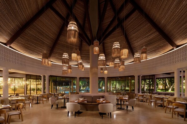Un espacio interior amplio con varias mesas y sillas para comer, una zona de estar central y ventanas grandes que ofrecen vistas al exterior.