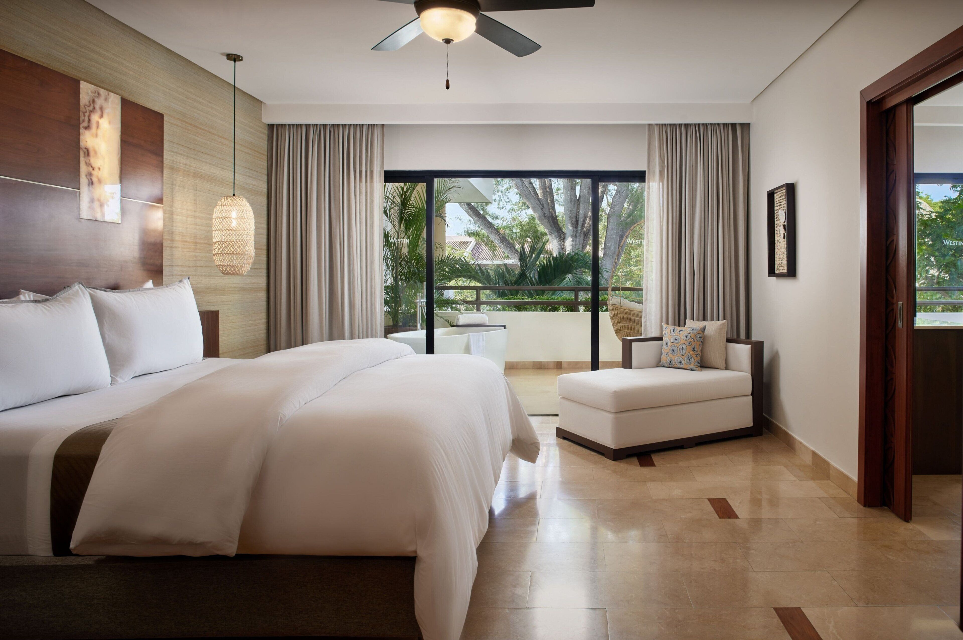 A modern hotel room with a large bed, a sofa, a ceiling fan, and a view of the outdoors.
