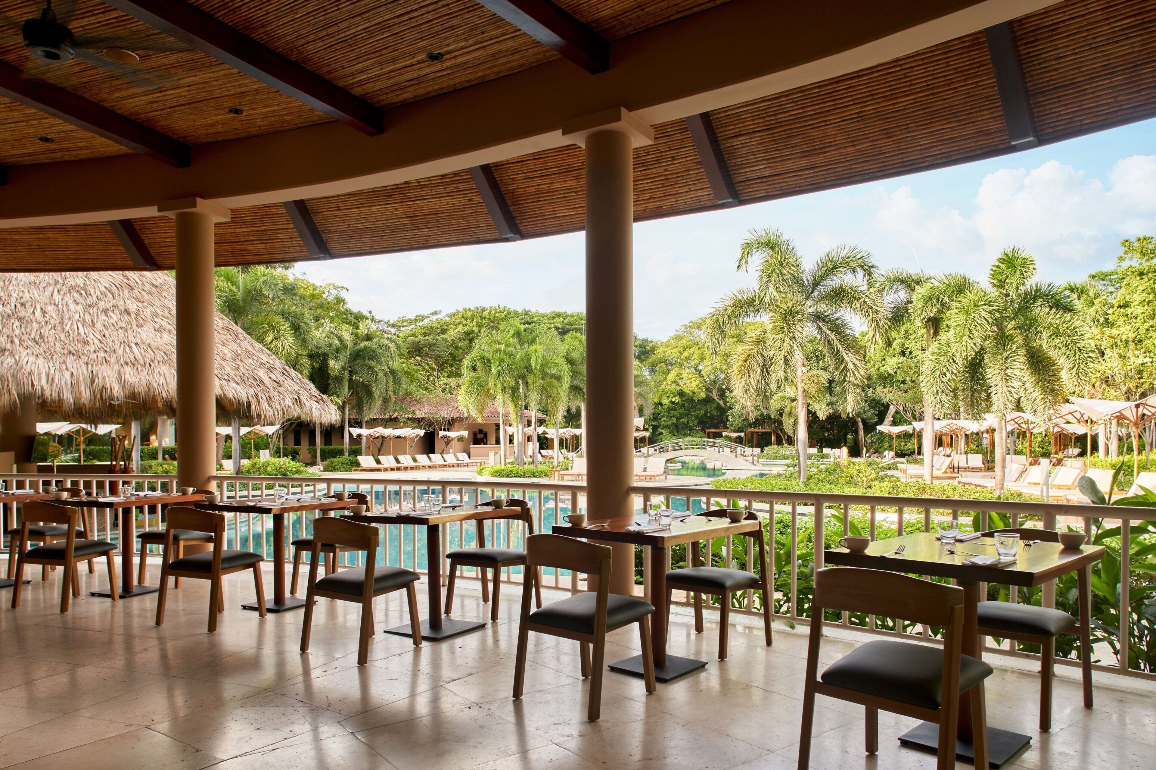 A covered outdoor dining area with tables and chairs, a pool, and lush greenery in the background.