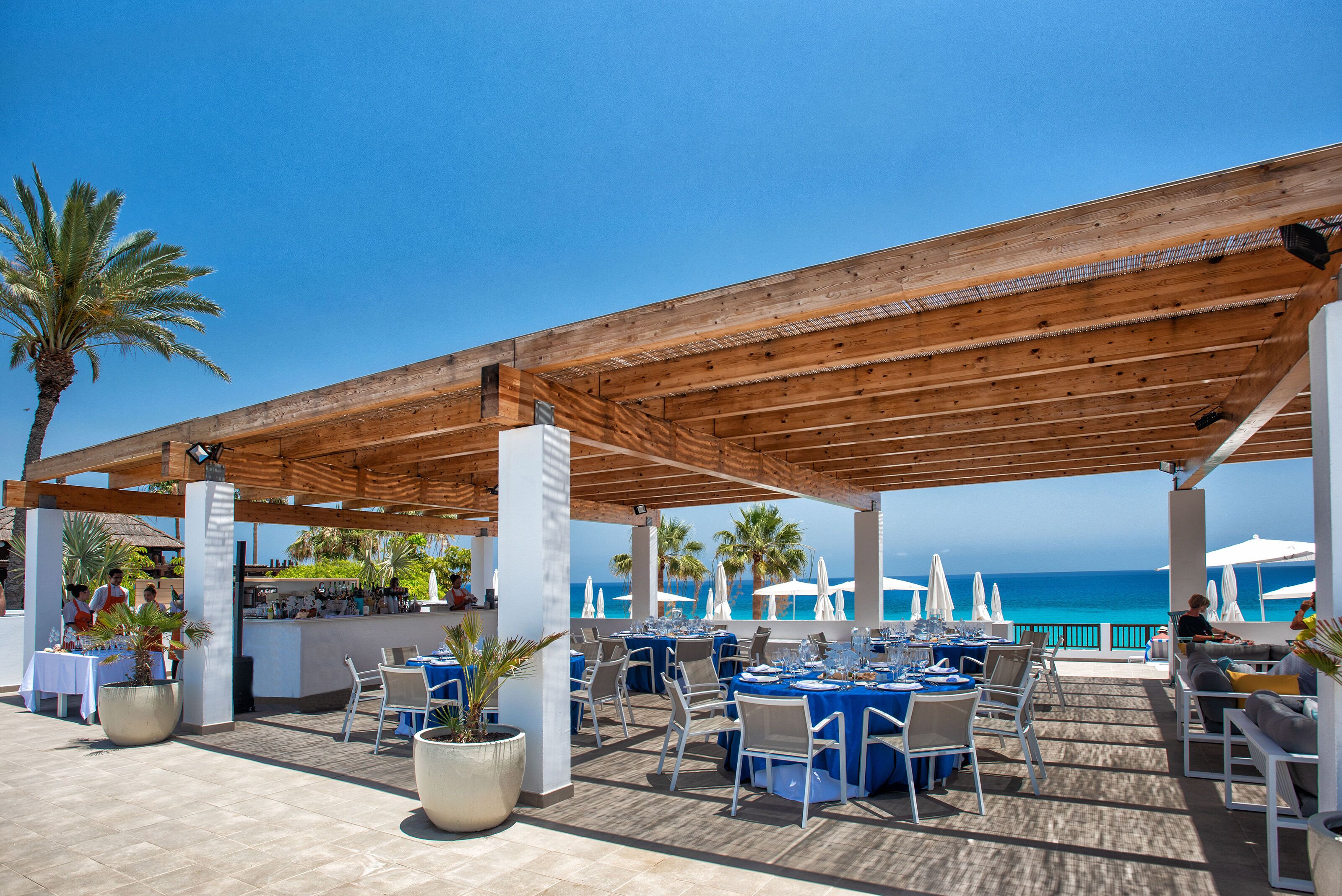 An outdoor dining area with tables and chairs, a wooden pergola, and a view of the ocean.