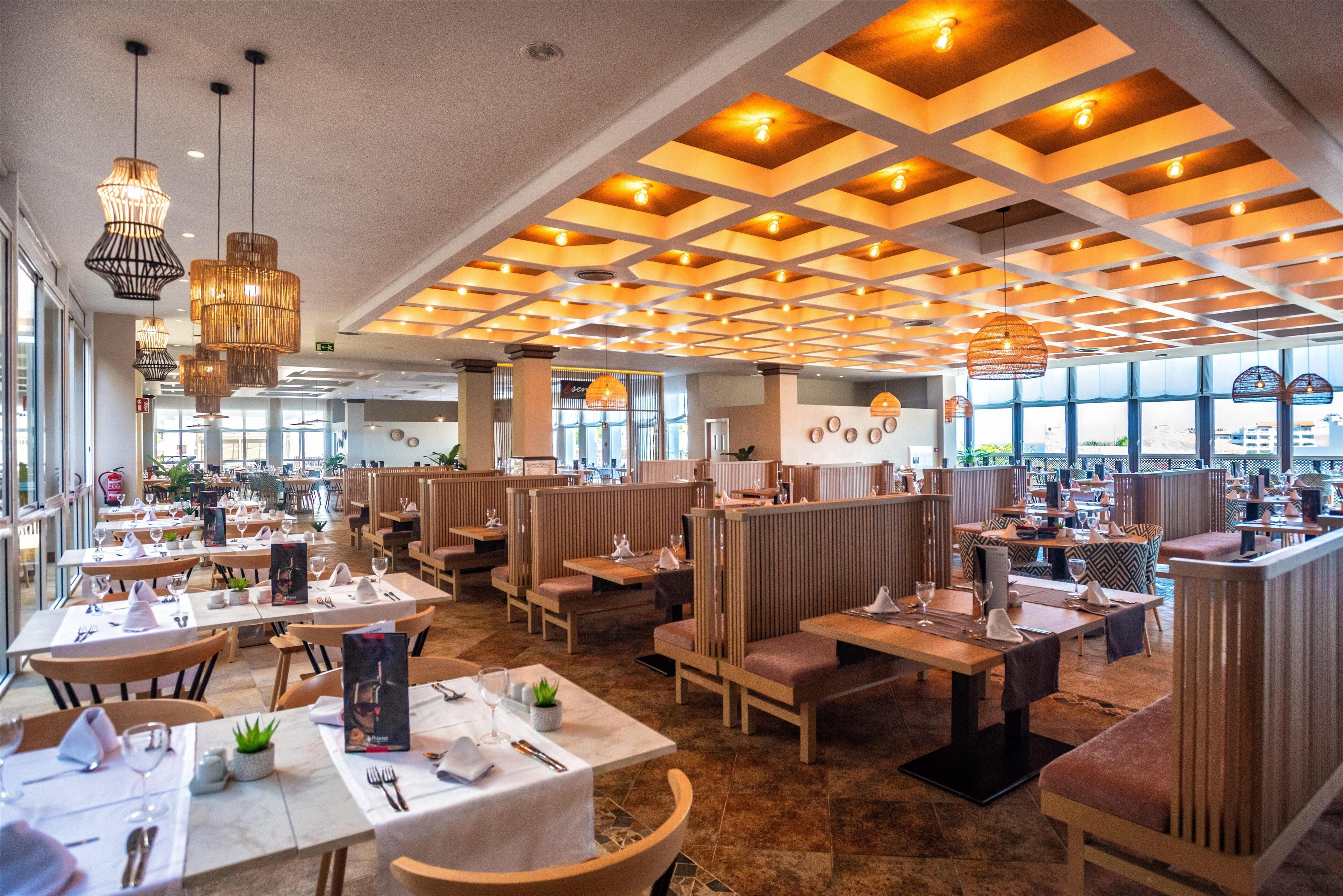Um restaurante espaçoso com pé direito alto, painéis de madeira e iluminação pendente moderna. As mesas estão organizadas com toalhas brancas e talheres postos.