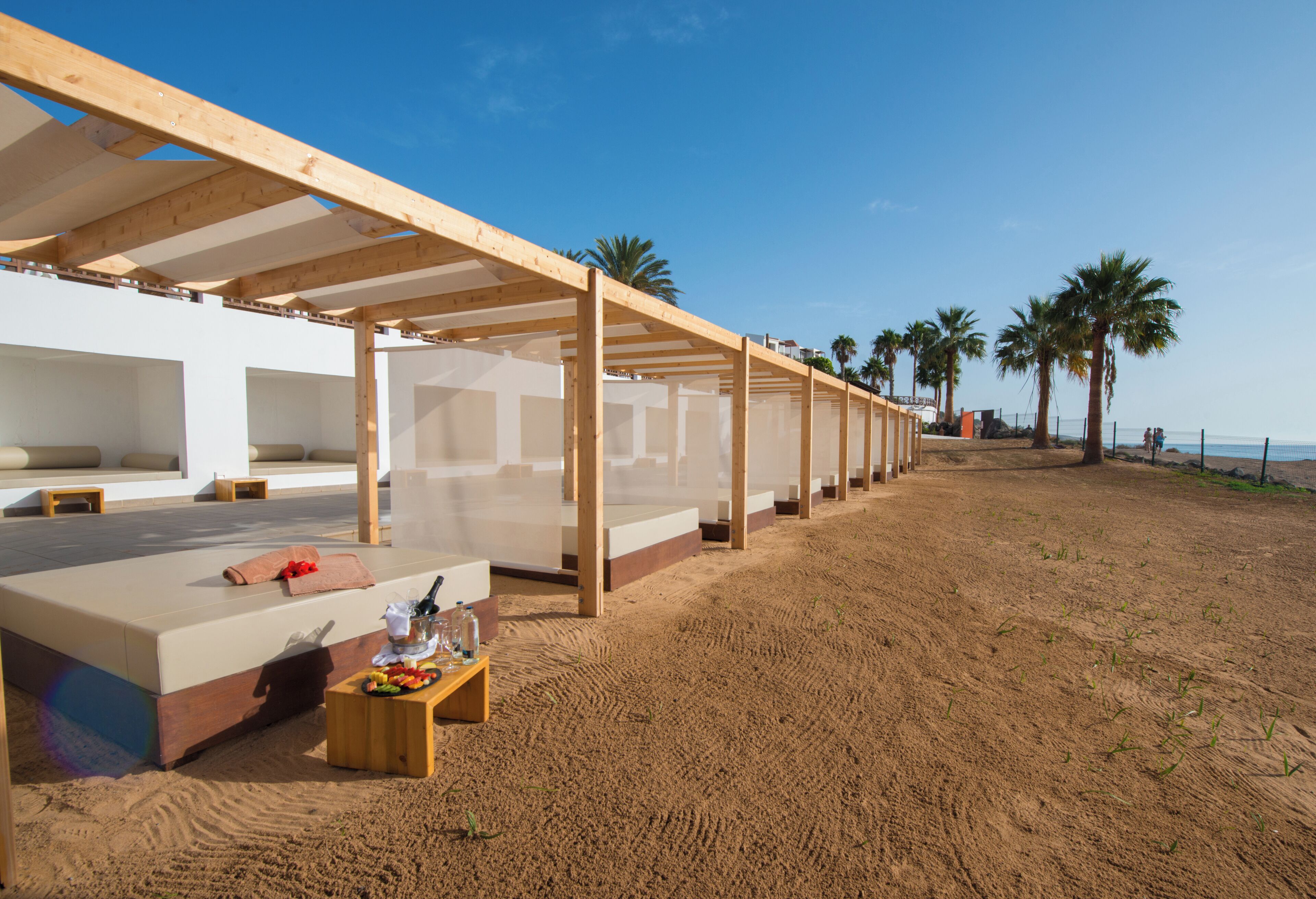 A beachside hotel with outdoor seating areas, a sandy floor, and palm trees in the background.
