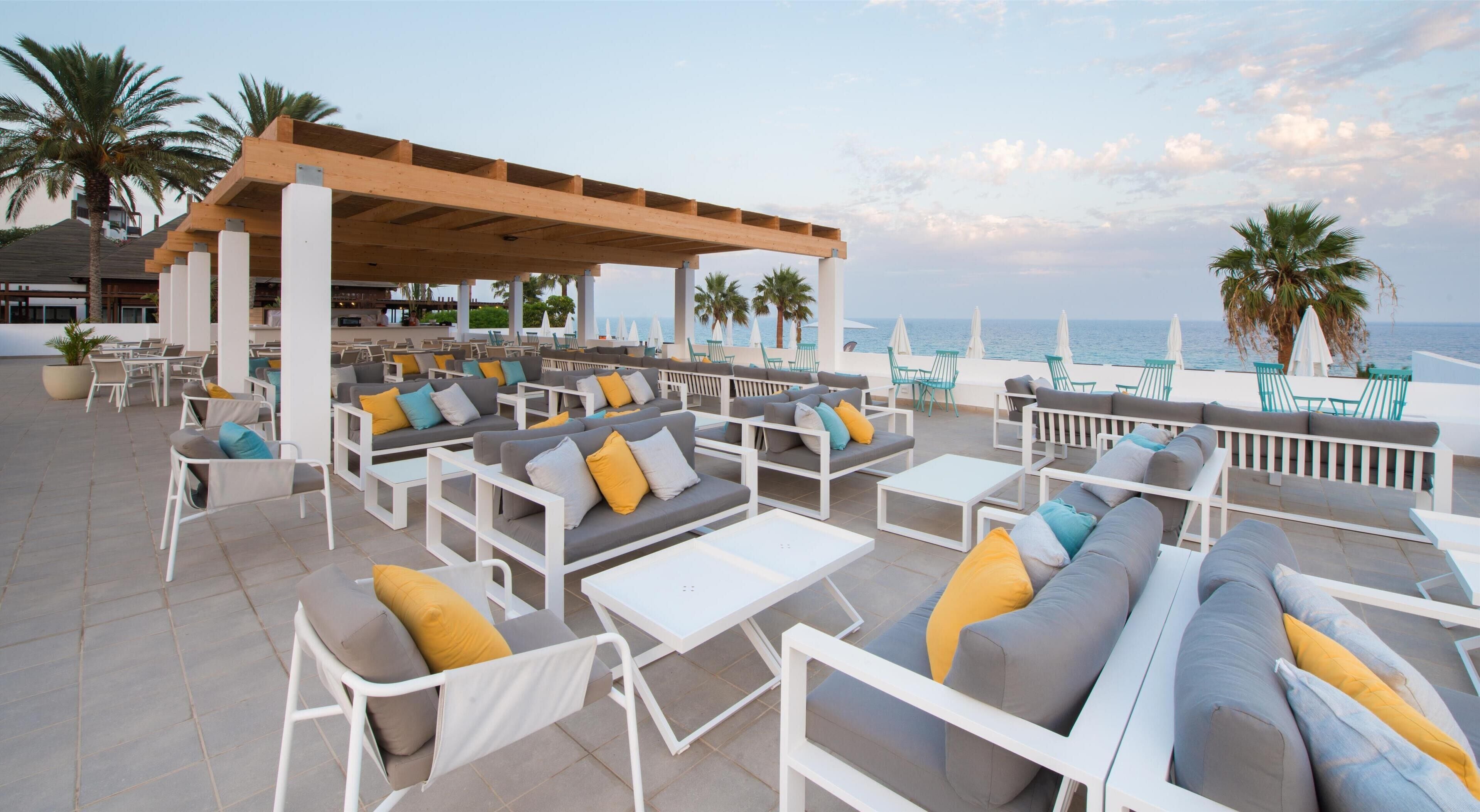 An outdoor seating area with multiple sofas and chairs, a wooden pergola, and a view of the sea.