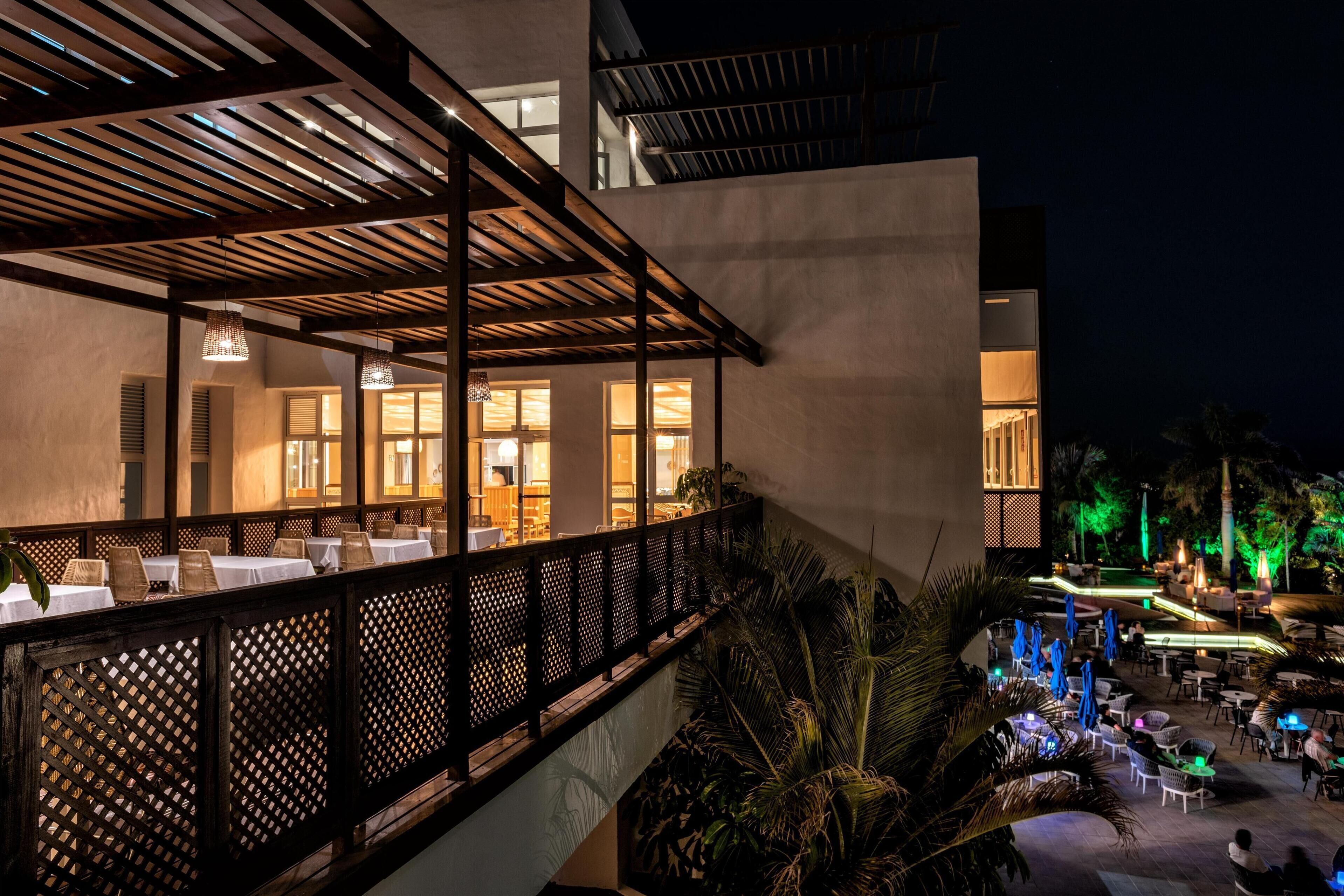 An outdoor dining area with tables and chairs, a wooden pergola, and a pool area with lights.