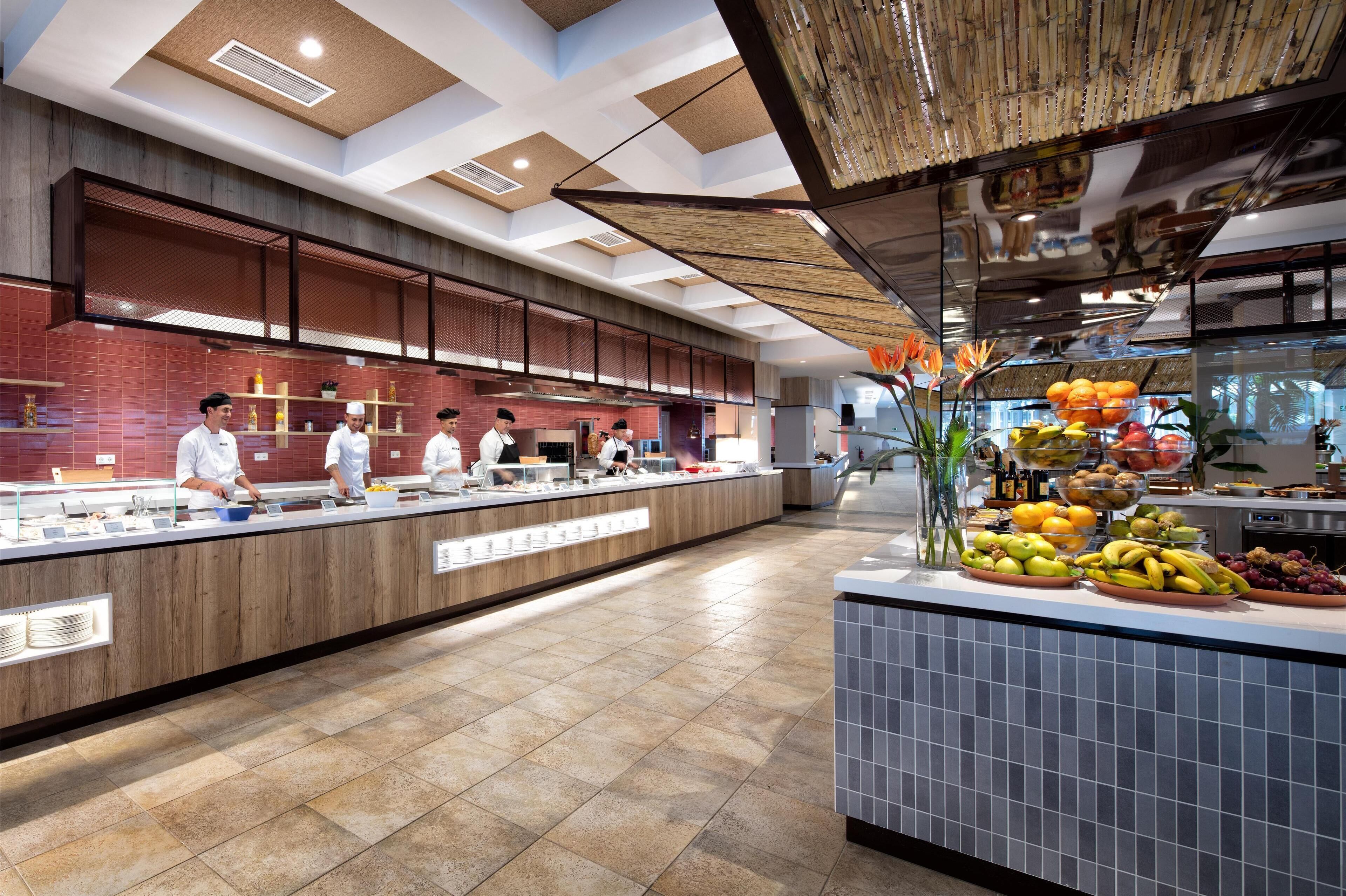 Een moderne keuken met koks die gerechten bereiden, een buffet met fruit en groenten, en een schone, betegelde vloer.