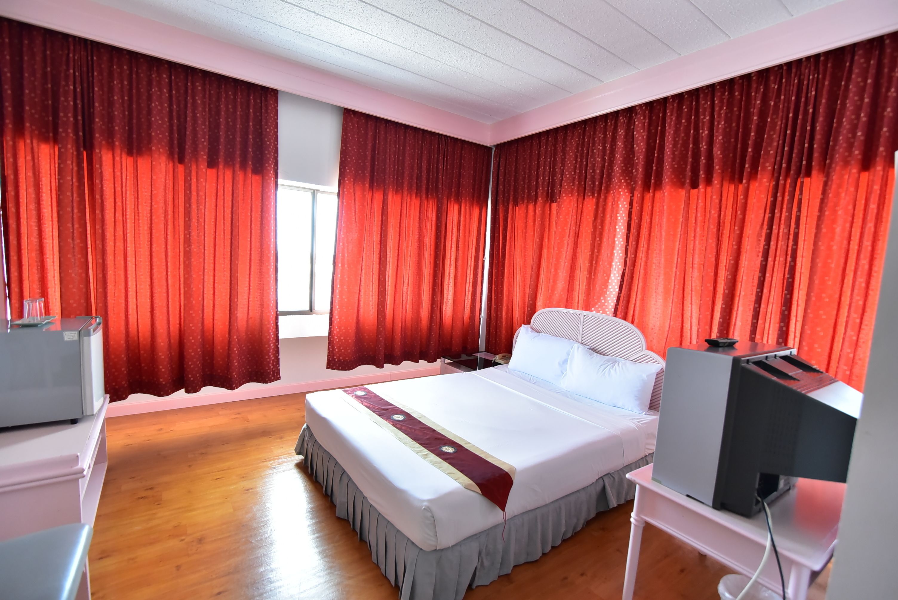 Quarto de hotel com cama, televisão sobre suporte e cortinas vermelhas.