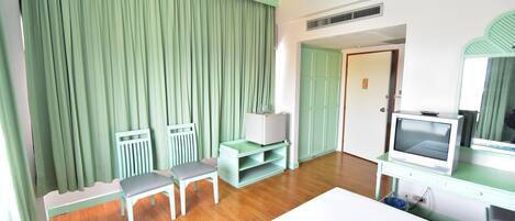 A room with a bed, a TV, a desk, and a chair. There is a door leading to another room.