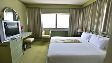 A hotel room with a bed, a desk with a television, a chair, a window with curtains, and a nightstand with a lamp.