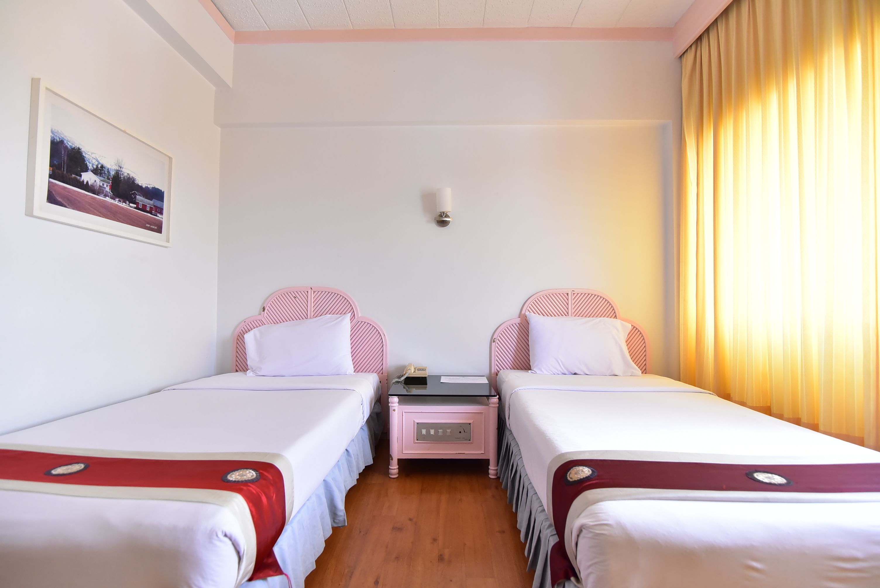 A hotel room with two beds, a nightstand, a wall-mounted light, and a window with curtains.