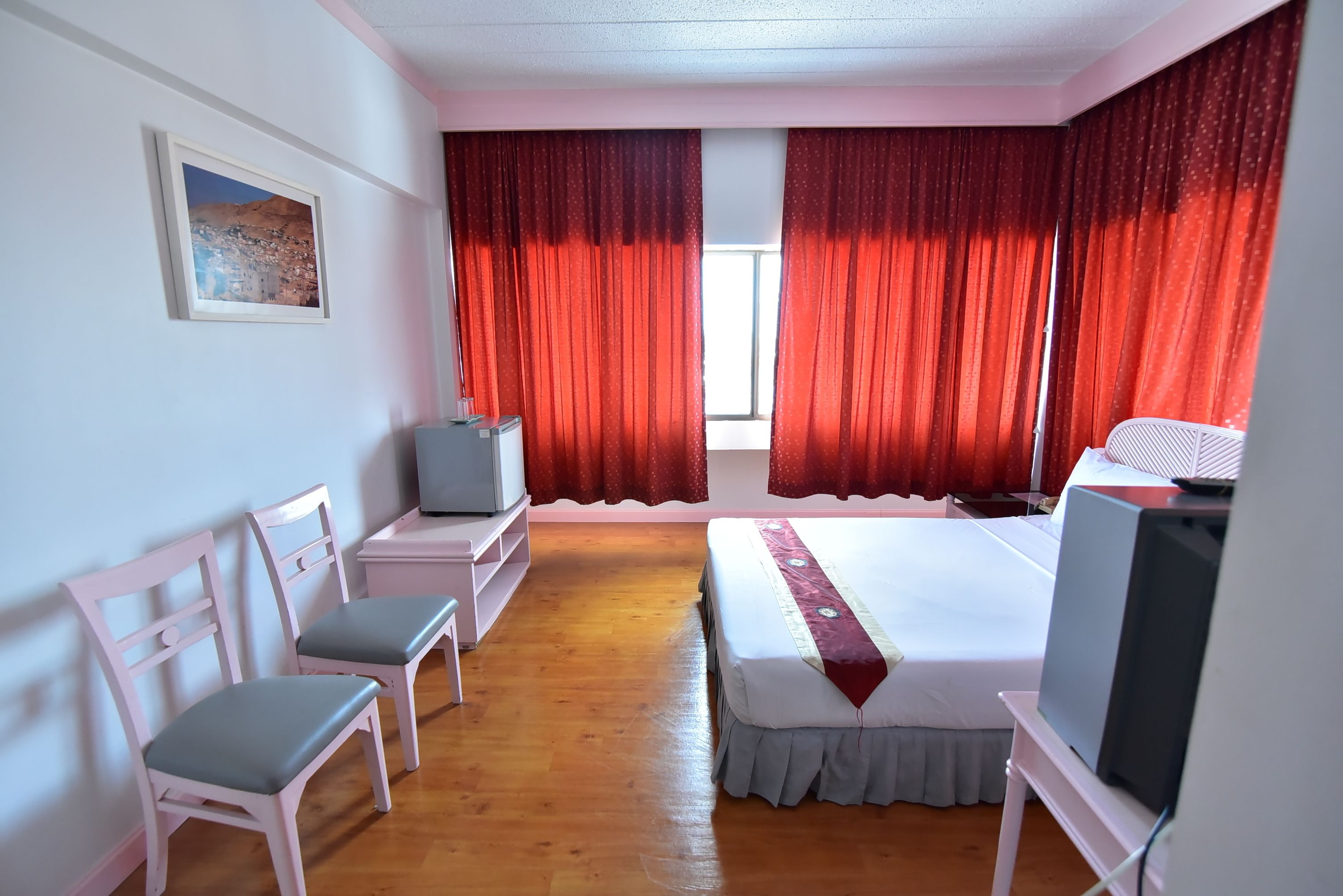 A hotel room with a bed, a television, a desk, two chairs, and a window with red curtains.