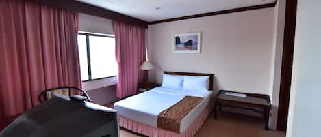 Quarto de hotel com cama, escrivaninha, cadeira, mesa de cabeceira e janela com cortinas.