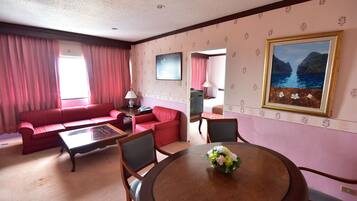 A hotel room with a red sofa, a glass-top table with chairs, a wooden dining table with a flower centerpiece, and a painting of a coastal scene.