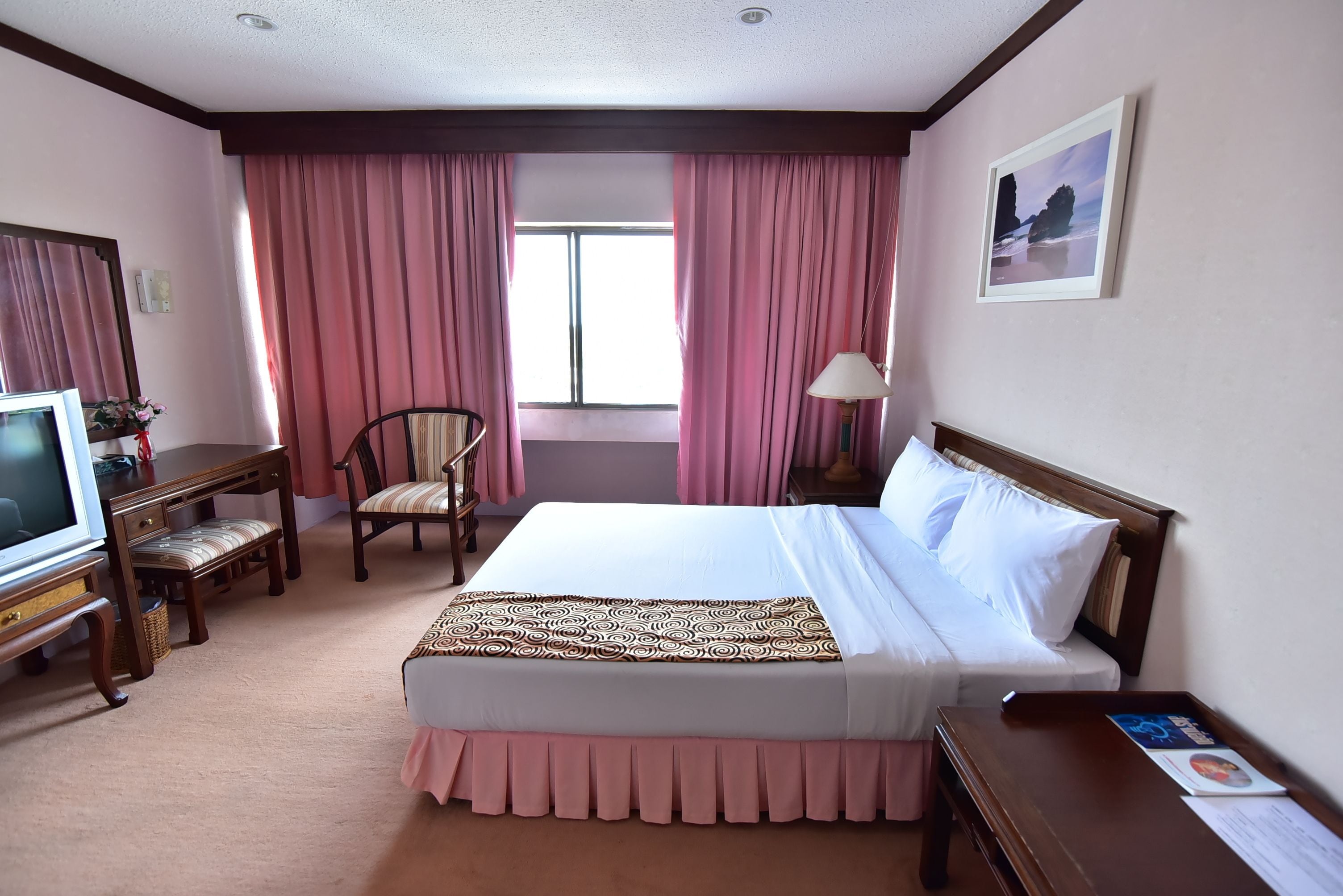A hotel room with a large bed, a desk with a television, a chair, and a window with pink curtains.