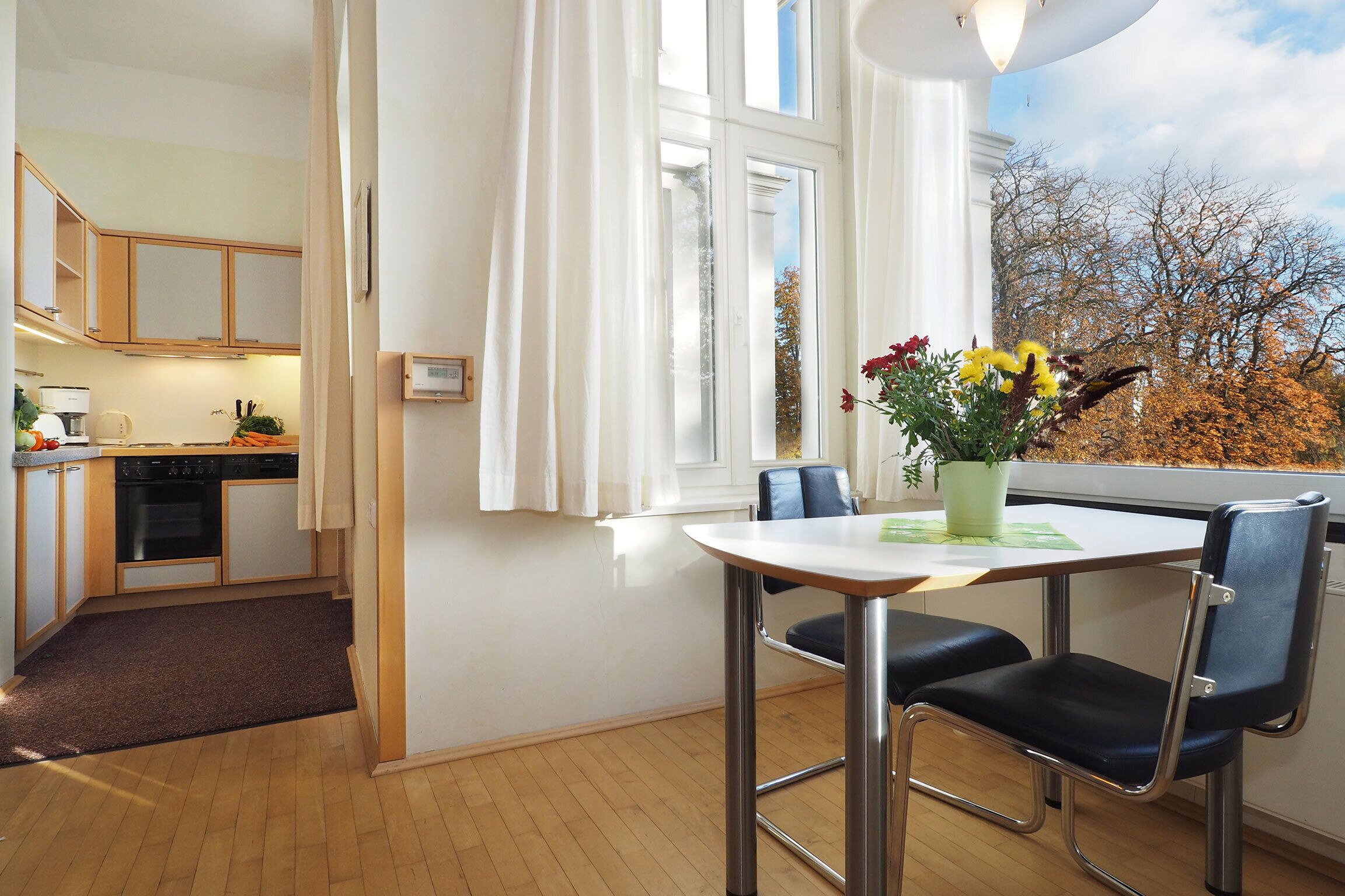 A modern kitchen with wooden cabinets, a dining area with a table and chairs, and a view of trees through a window.