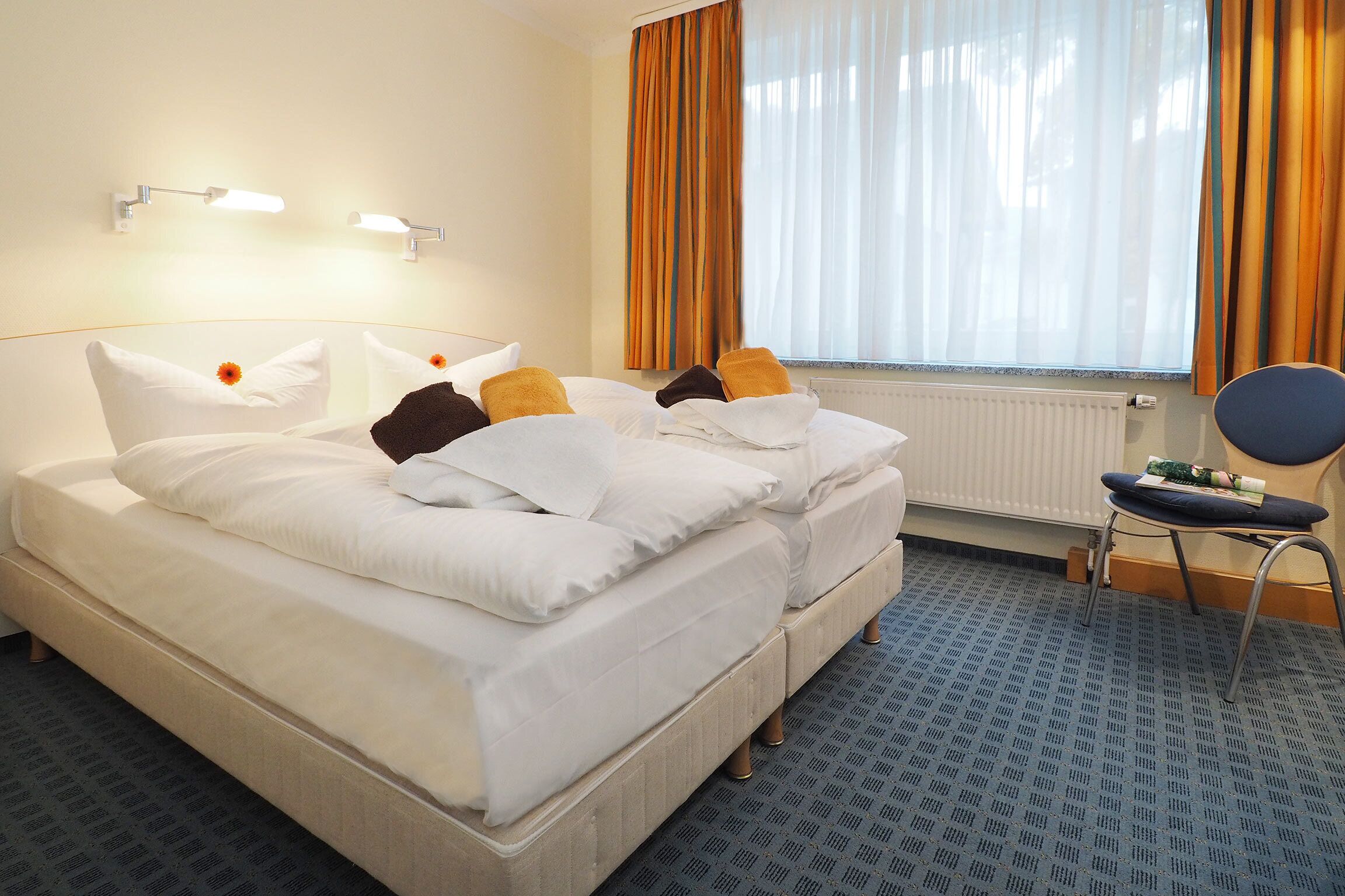 Habitación de hotel con cama doble, dos mesitas de noche, una silla, una ventana con cortinas y un radiador.