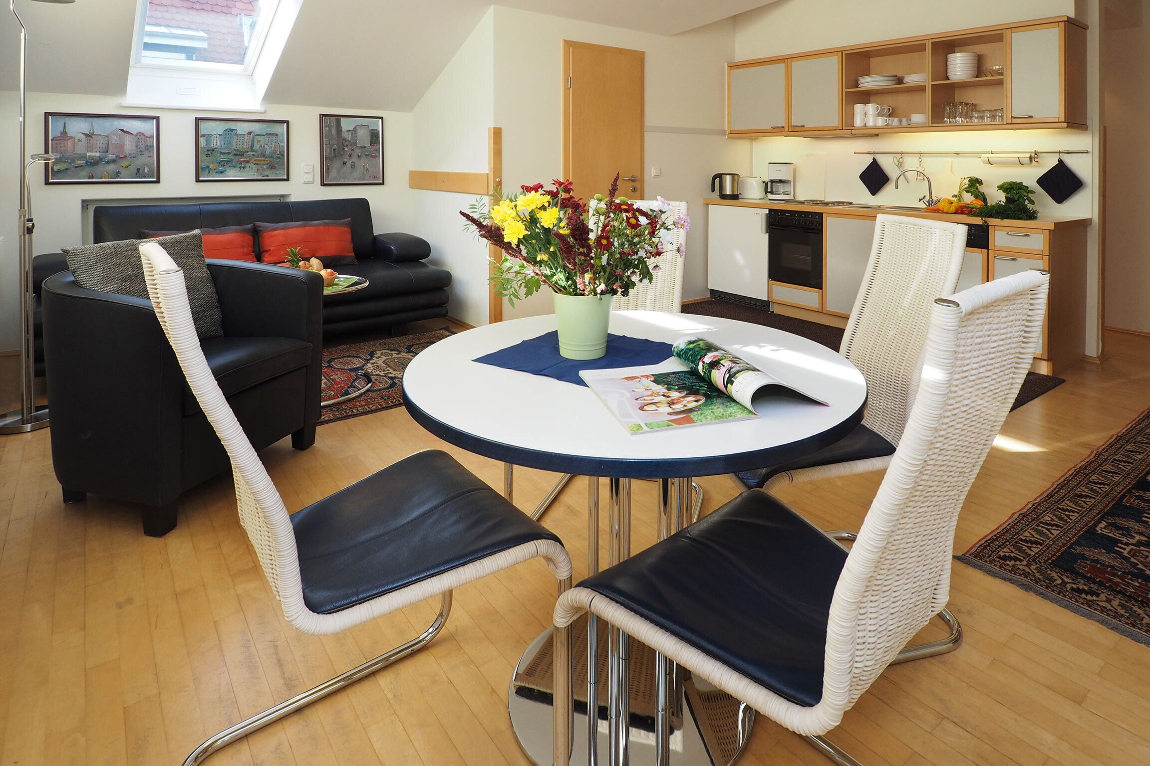 Ein modernes Wohnzimmer mit einem schwarzen Sofa, weißen Korbstühlen, einem runden Tisch mit Blumen und einer Küche im Hintergrund.