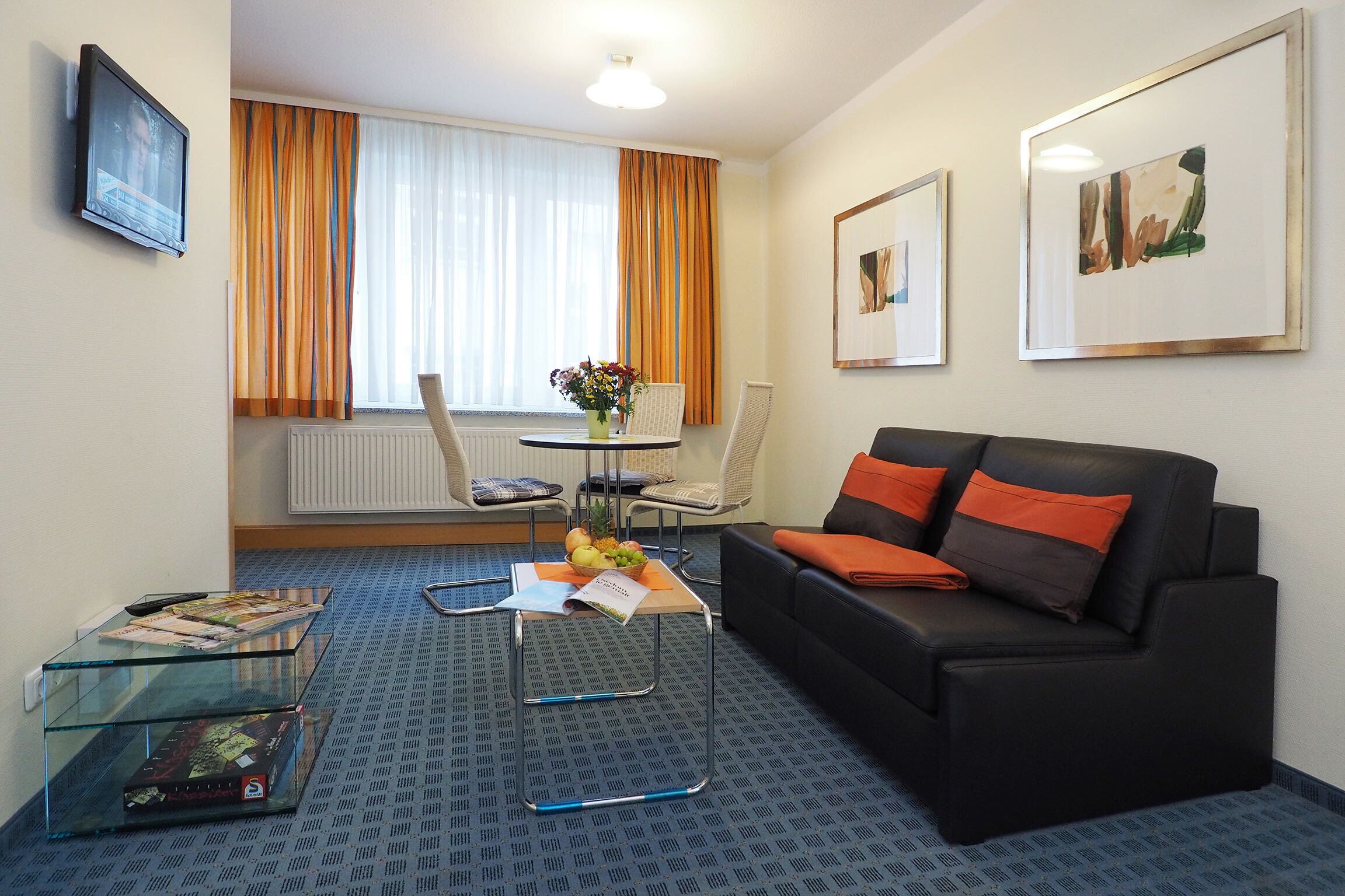 Ein modernes Hotelzimmer mit einer schwarzen Ledersofa, einem Glastisch, einem kleinen runden Tisch mit Blumen und einem an der Wand montierten Fernseher.
