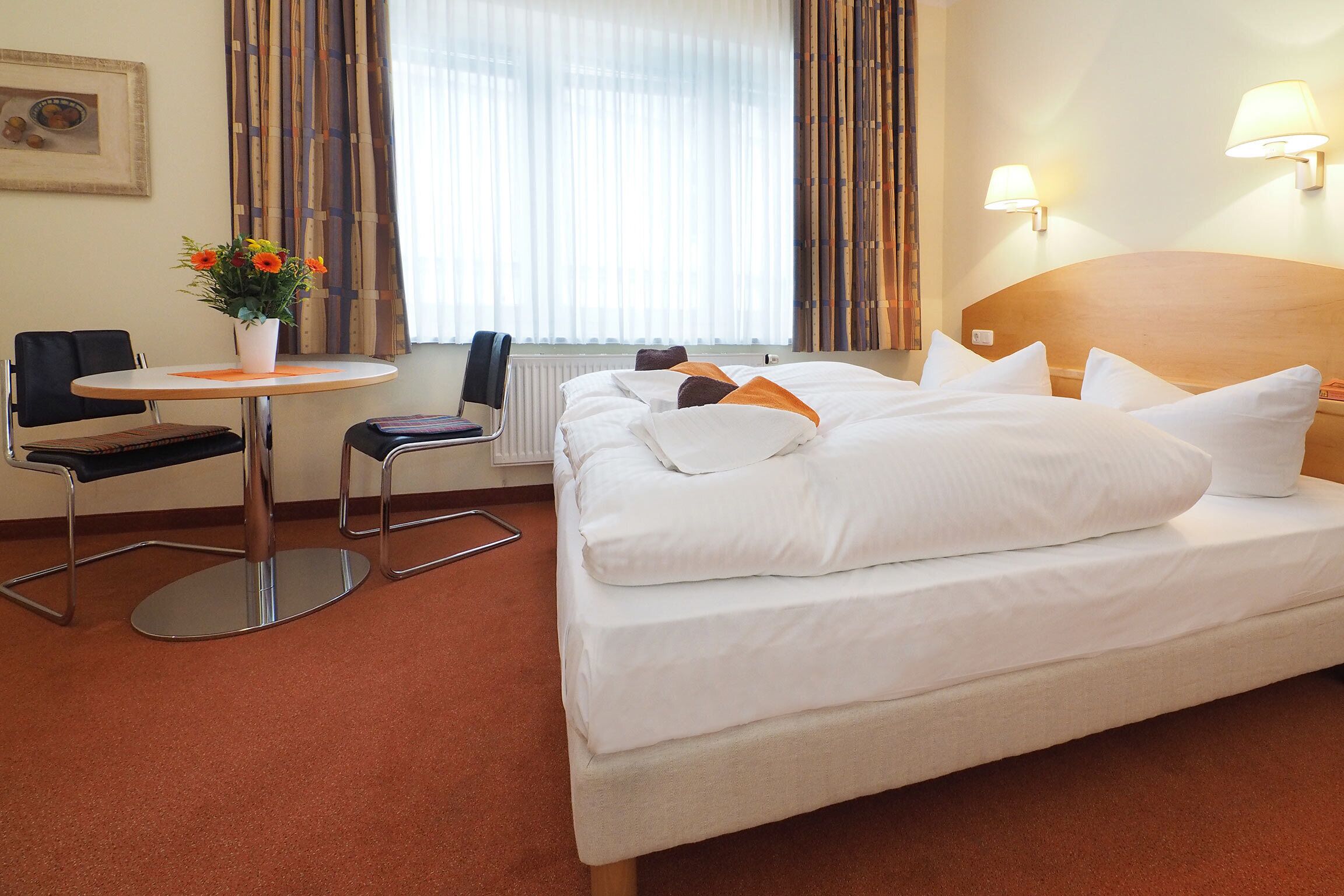 A hotel room with a bed, a small table with a vase of flowers, a chair, and a window with curtains.