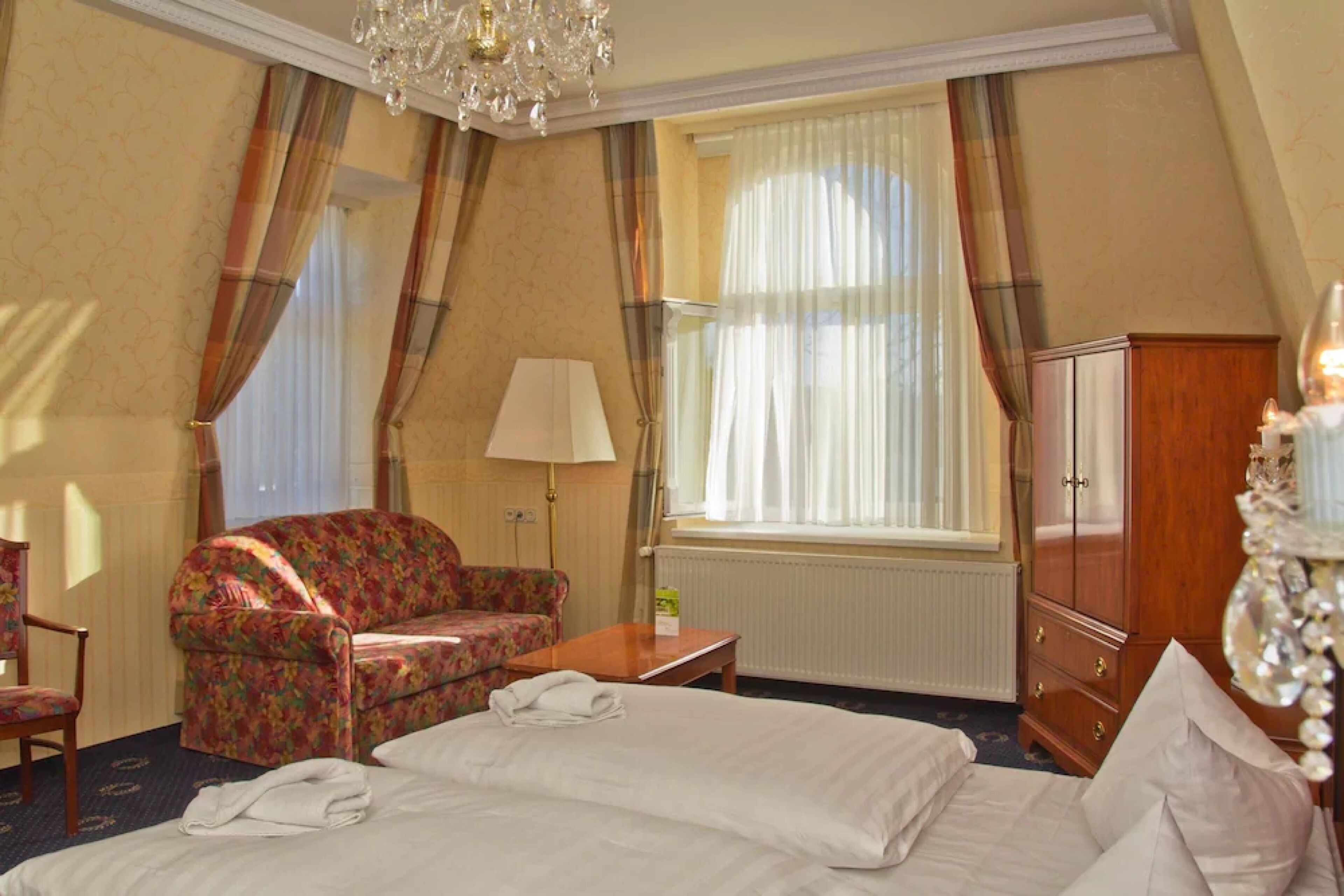 Habitación de hotel con cama, una silla, una mesa, lámpara, armario y ventana con cortinas.