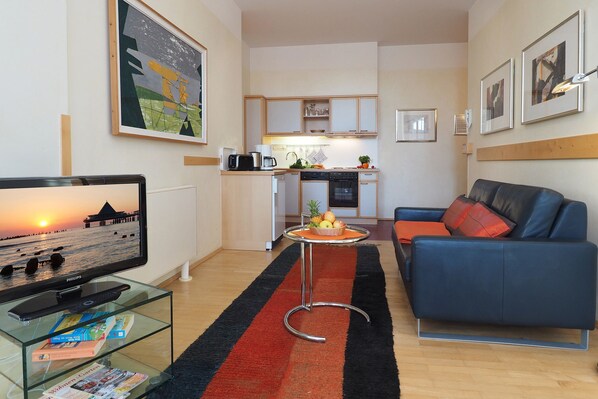 Una sala de estar moderna con televisor, una mesa de centro de vidrio y un sofá azul. Se ve una cocina al fondo con un comedor.