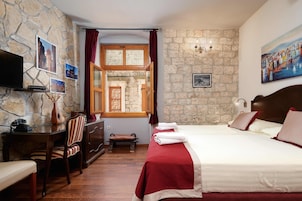 A hotel room with a stone wall, a bed with red bedding, a desk with a telephone, a chair, and a window with curtains.