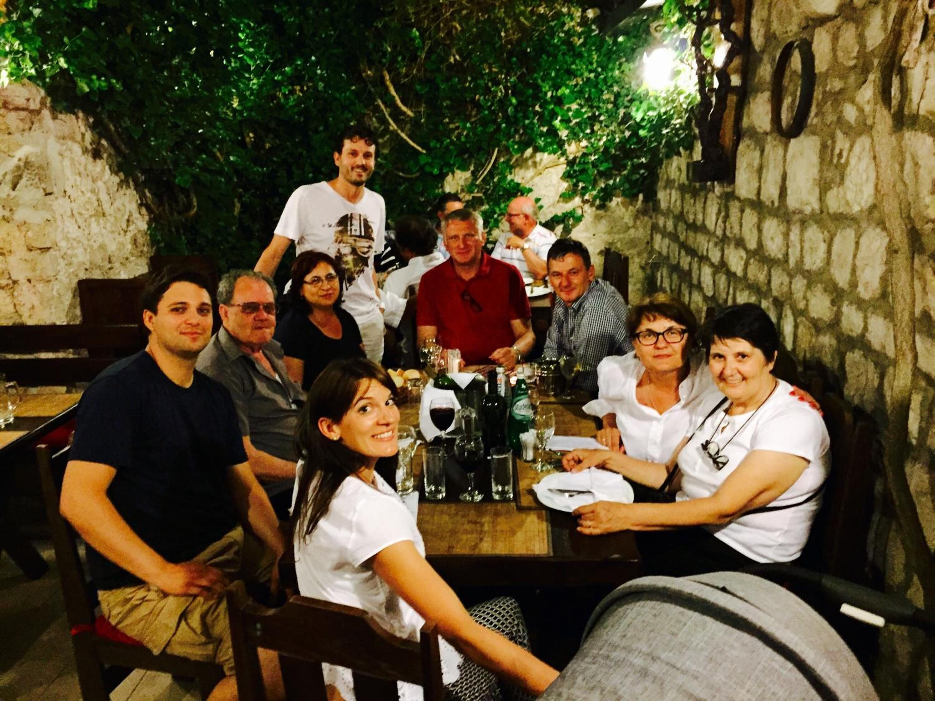 Un grupo de personas sentadas alrededor de una mesa en un espacio al aire libre, con paredes de piedra y follaje.