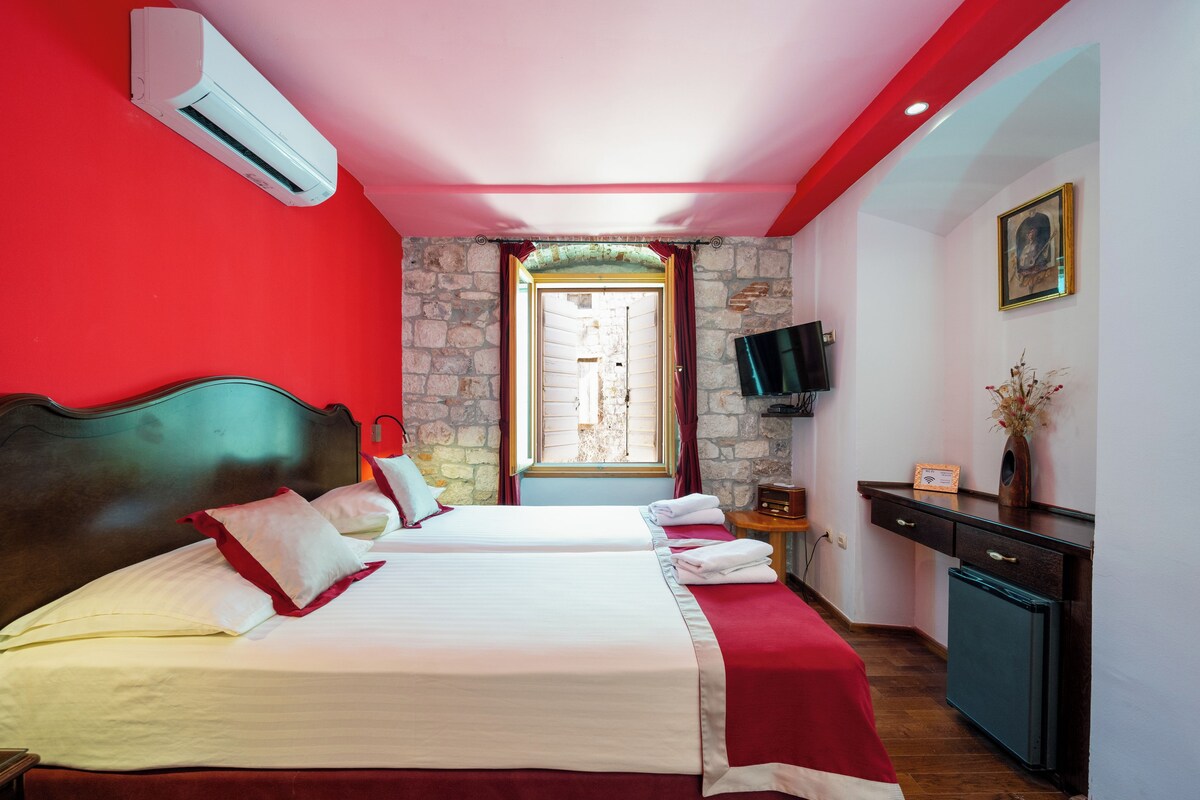 A hotel room with two beds, a stone wall, a TV, and a desk with a vase.
