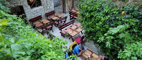 An outdoor patio area with wooden tables and benches, surrounded by lush greenery and stone walls.