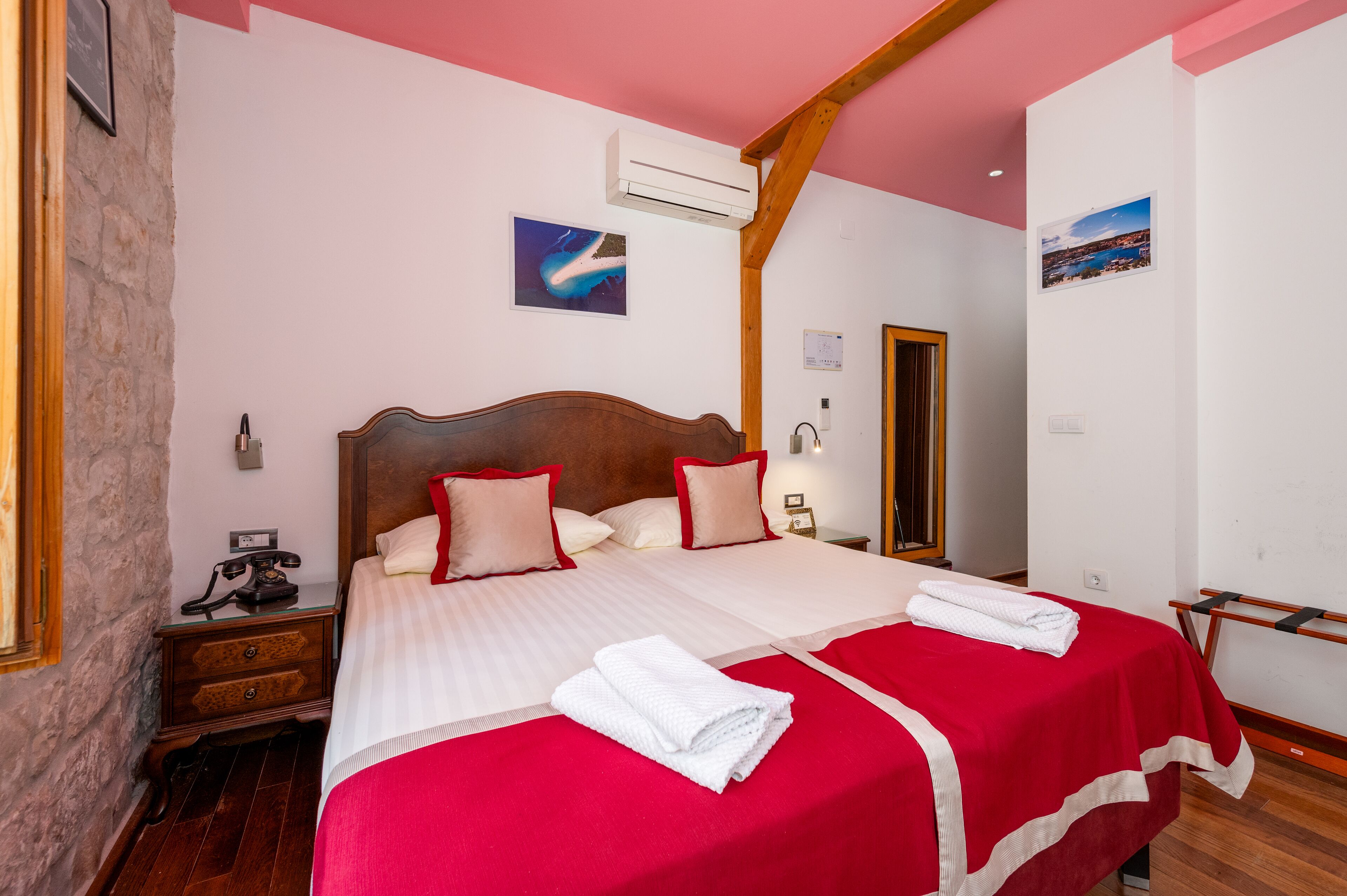 A hotel room with a large bed, wooden headboard, and a red bedspread. There is a wooden nightstand with a telephone and a small table with a lamp. The room has white walls and a stone wall on one side.