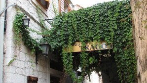 Un estrecho callejón adoquinado con un restaurante llamado "TRAGOS" bajo un dosel de hiedra.