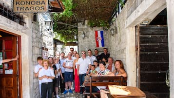 A group of people gathered in a narrow street with stone walls and a wooden bench. A sign above reads "KONOBA TRAGOS."