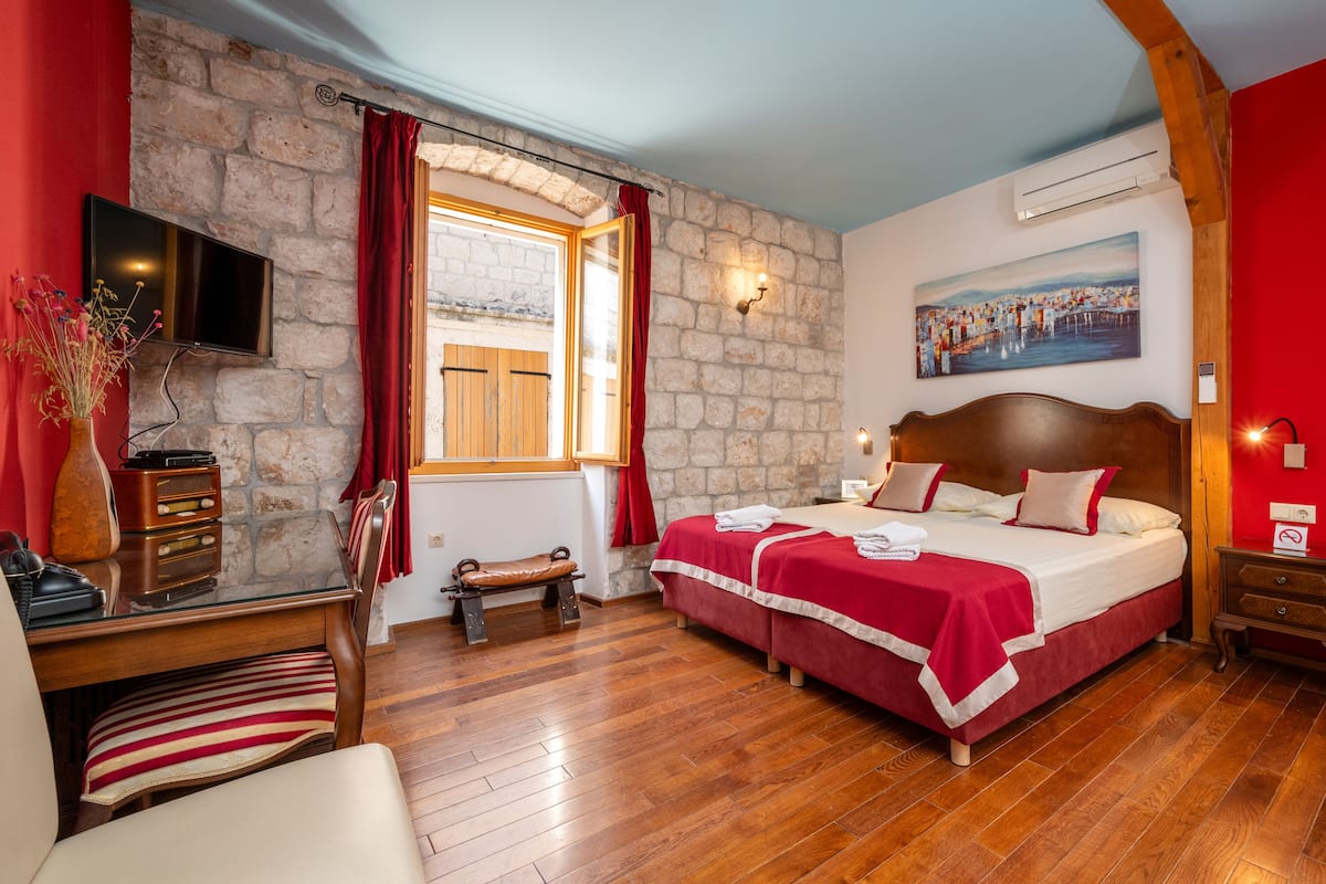 A hotel room with a stone wall, a bed with red bedding, a wooden desk, a chair, a television, and a painting on the wall.