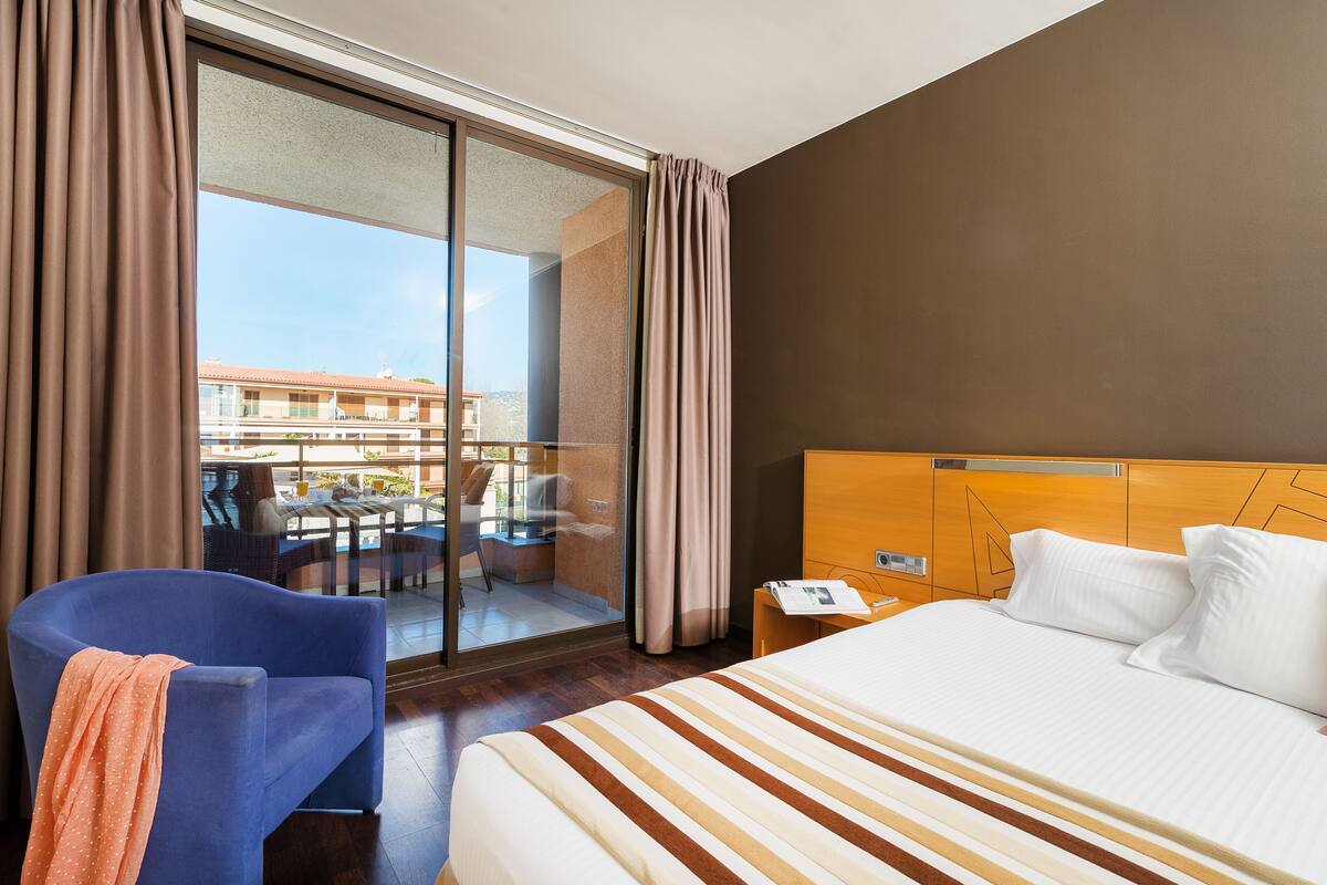Habitación de hotel con cama, silla, mesita de noche y vistas al balcón exterior.