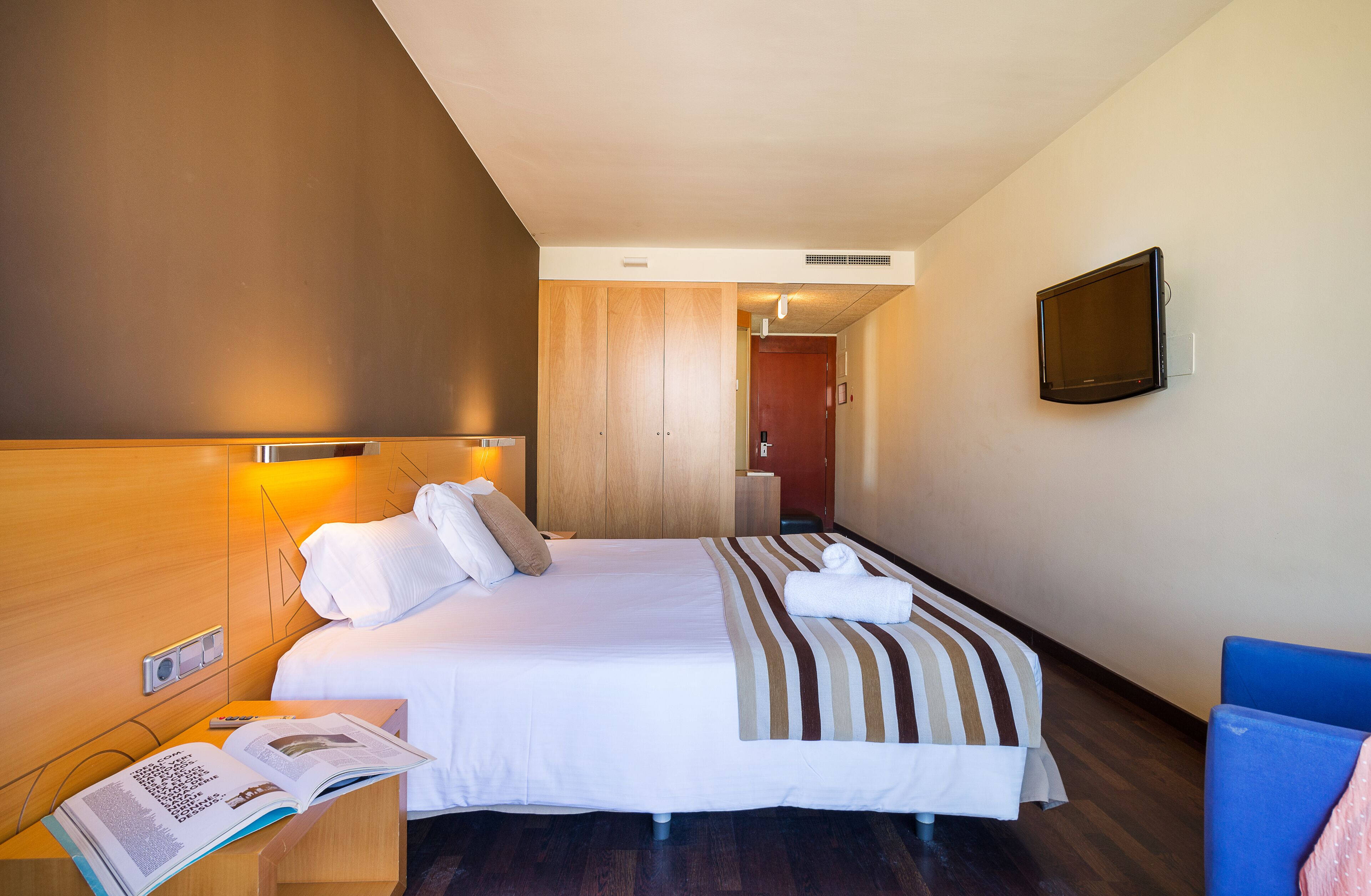 A hotel room with a bed, bedside table with a book, a TV on the wall, and a wooden wardrobe.