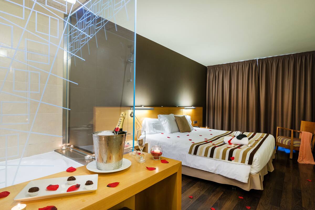 Habitación de hotel con cama, mesita de noche con una botella de champán, una copa de vino y pétalos de rosa en el suelo.