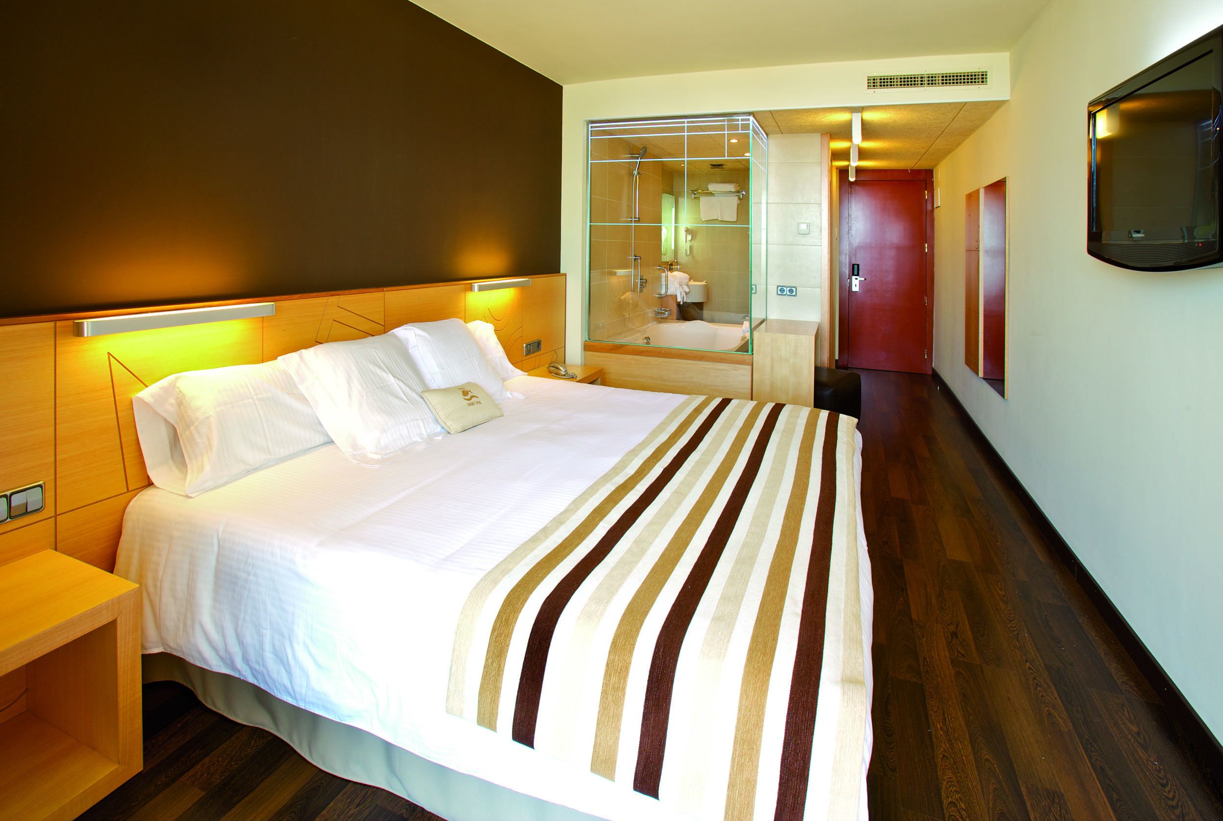 A hotel room with a large bed, a bedside table, a glass-enclosed shower, a mirror, and a TV mounted on the wall.