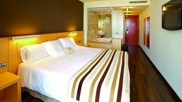 A hotel room with a large bed, a bedside table, a glass-enclosed shower, a mirror, and a TV mounted on the wall.