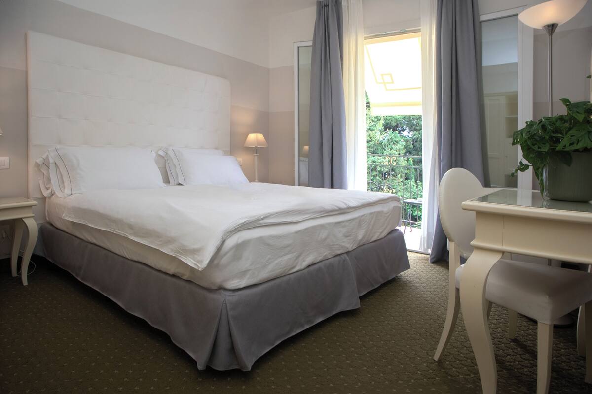 Ein Hotelzimmer mit einem großen Bett, einem Schreibtisch mit Stuhl, einem Blumentopf und Blick auf Grünfläche.