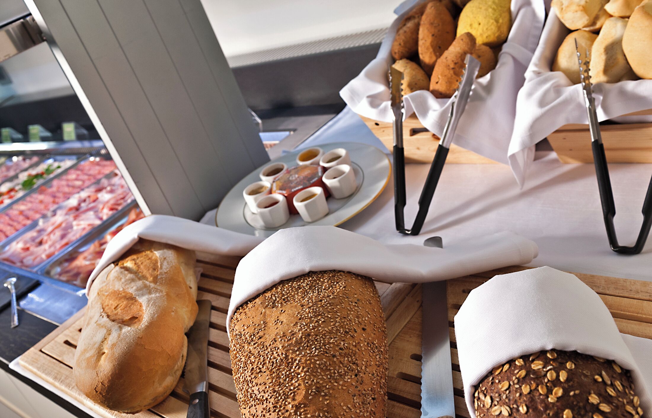 Diversos tipos de pães e doces sobre uma mesa de madeira, com um balcão de carnes ao fundo.