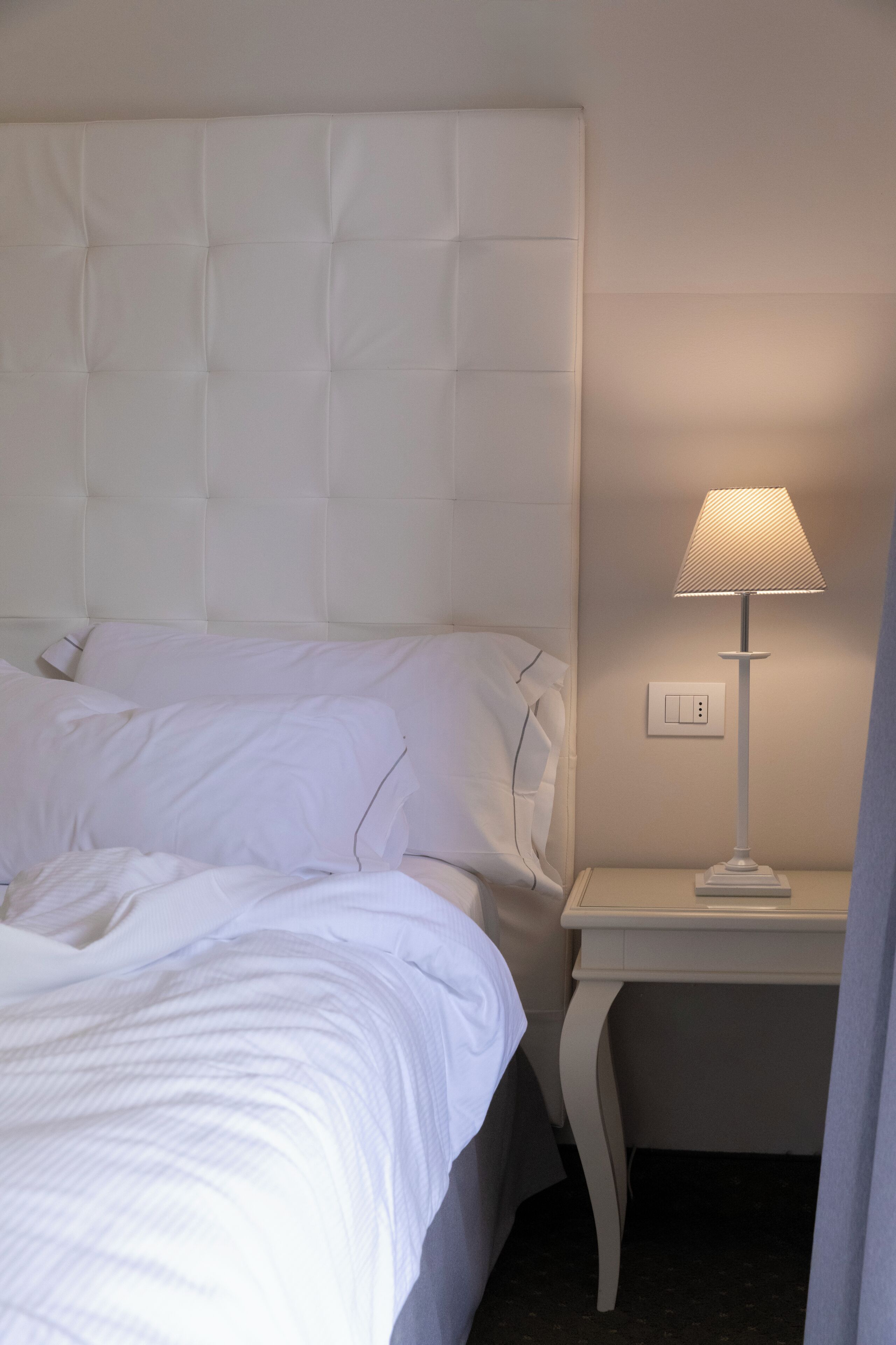 A neatly made bed with white linens, a white headboard, and a bedside table with a lamp.