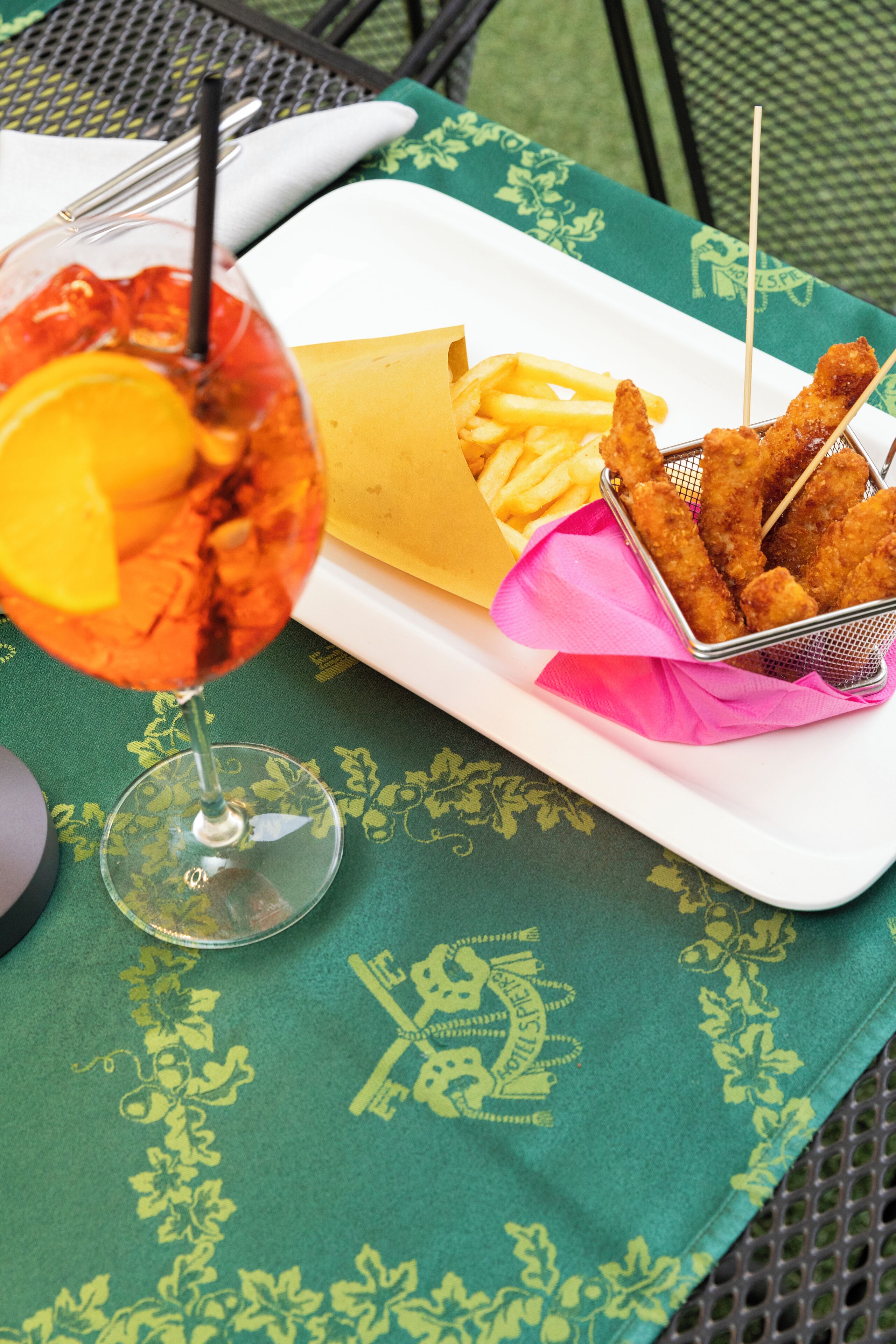 A meal consisting of a cocktail with a lemon slice, French fries, and breaded chicken tenders on a white plate, placed on a green tablecloth with a floral pattern.