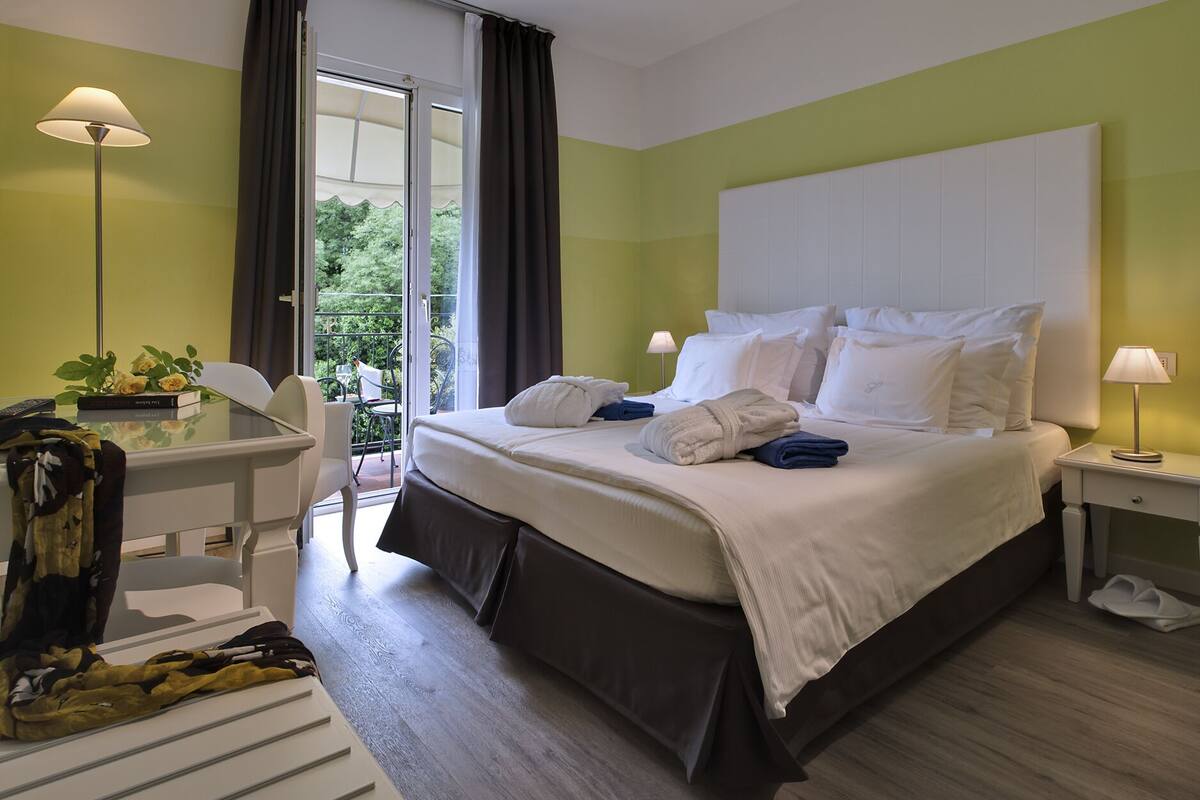 Ein Hotelzimmer mit einem großen Bett, zwei Nachttischen, einem Schreibtisch mit Stuhl und einem Balkon mit Blick auf Grünflächen.