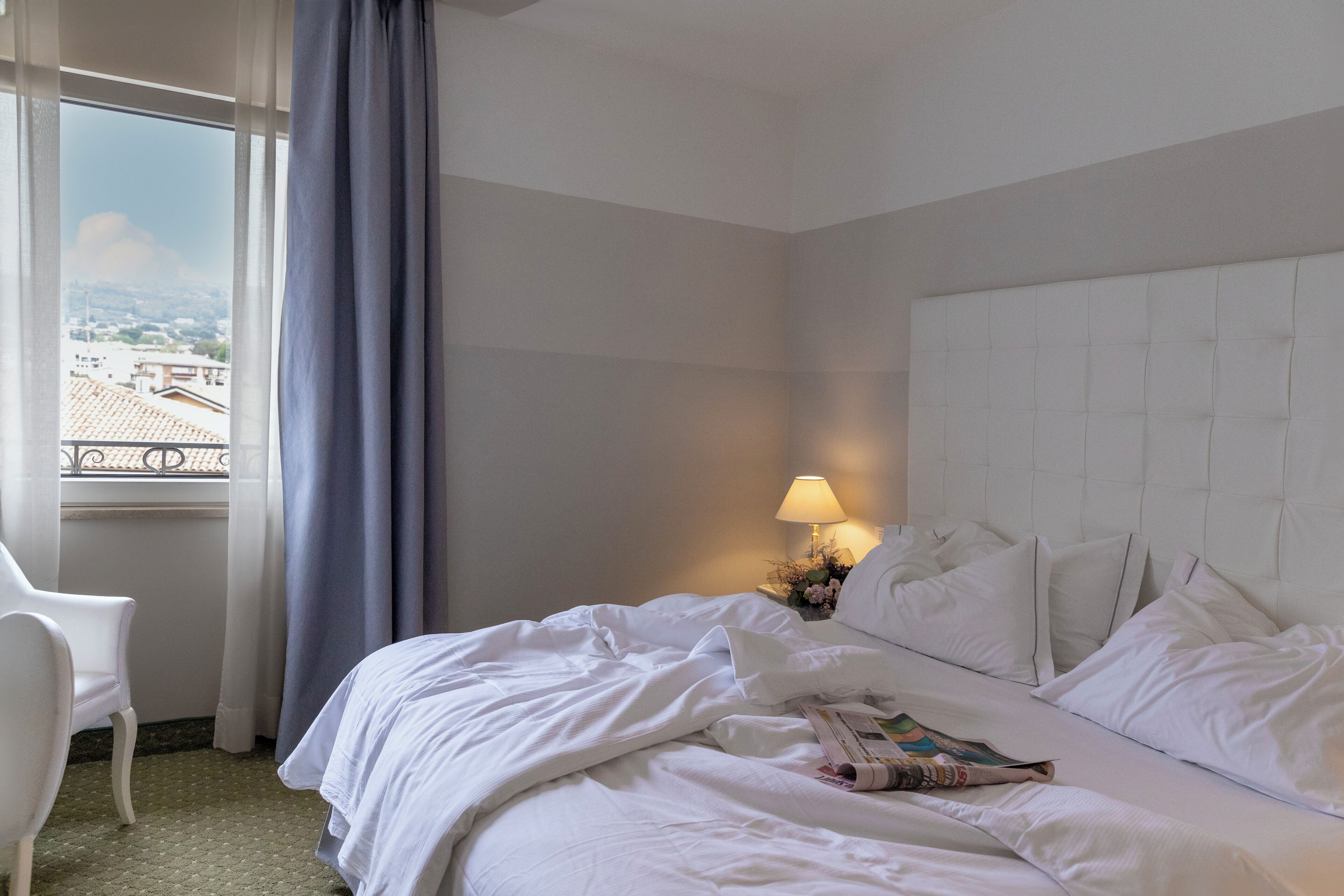 A hotel room with a bed, a chair, a nightstand with a lamp, and a view of the city through a window.