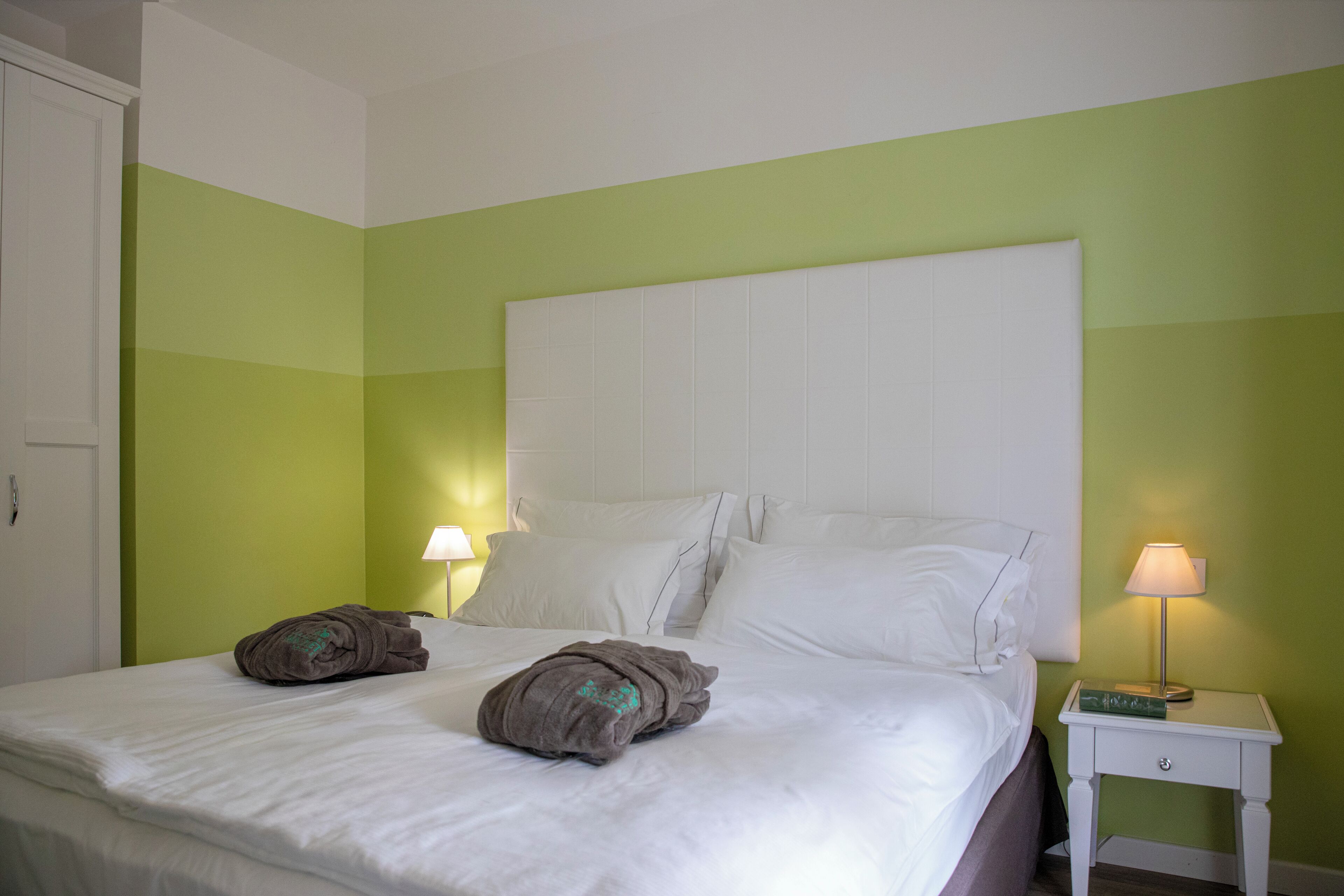 Een net opgemaakt bed met witte lakens en twee bruine handdoeken, een wit nachtkastje met een lamp, en een groene muur.