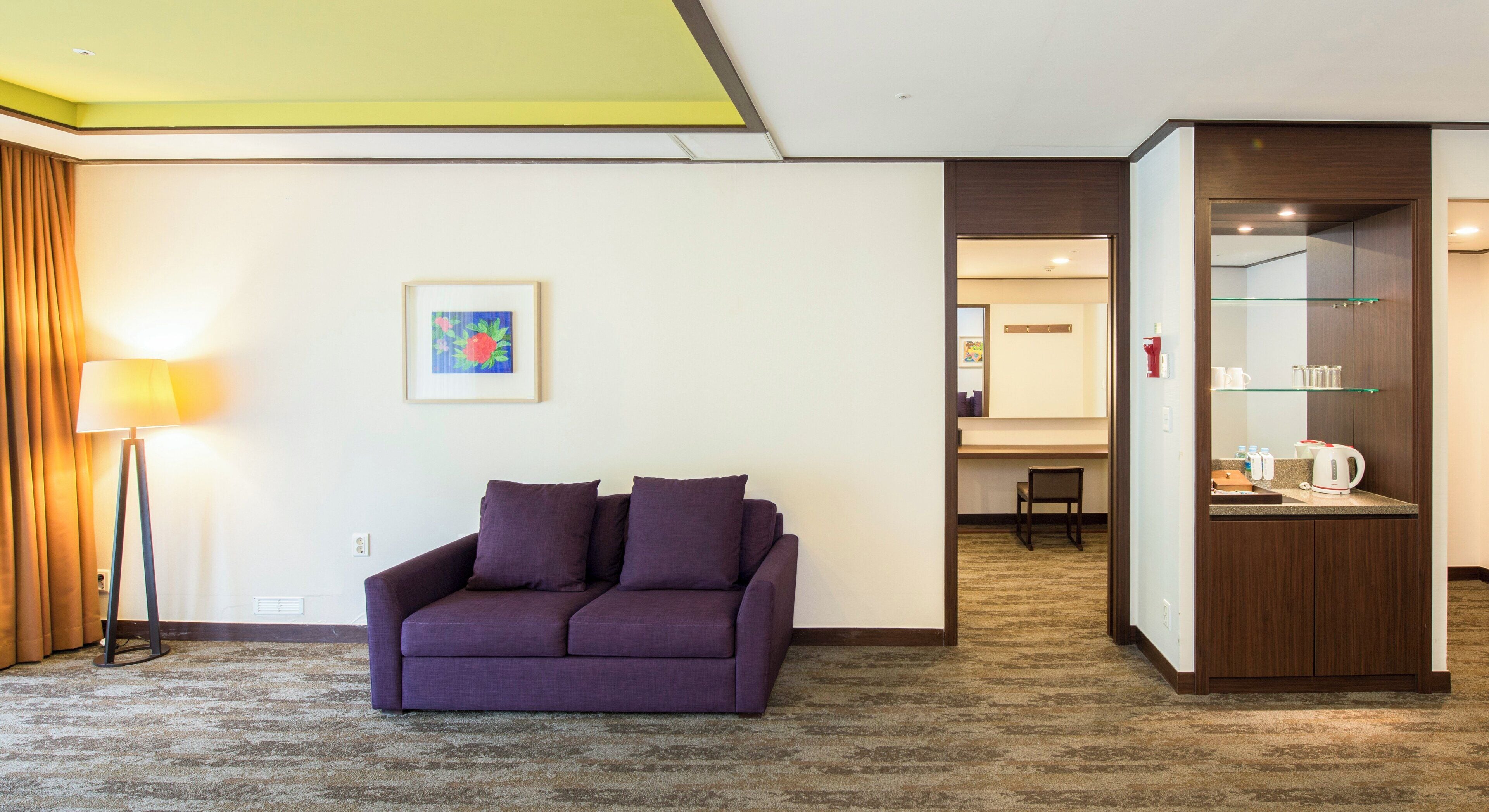 A hotel room with a purple sofa, a lamp, a framed picture, and a kitchenette area.