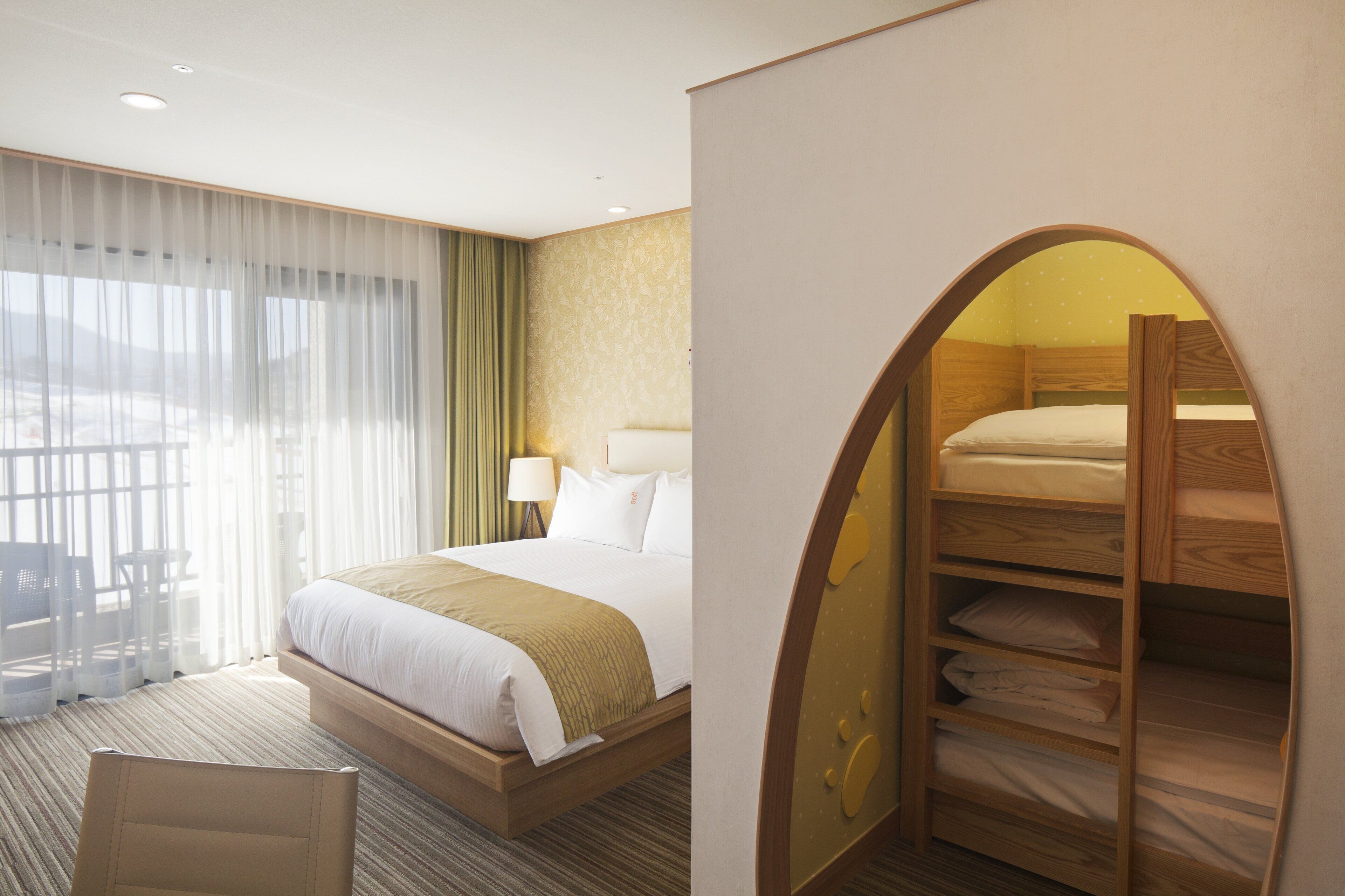 Una habitación de hotel con una cama, una zona de estar con una silla, una cama nido de madera y vistas a través de una ventana con cortinas traslúcidas.